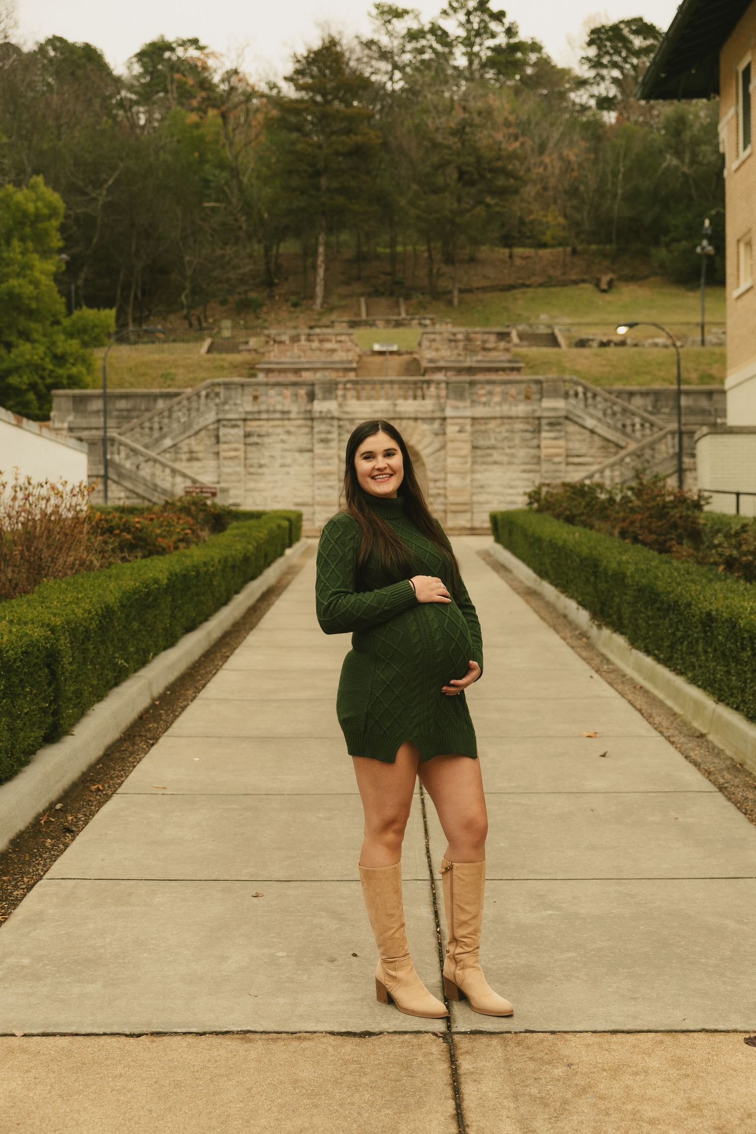 Smiling pregnant woman in green knit dress and tan boots standing in formal garden.