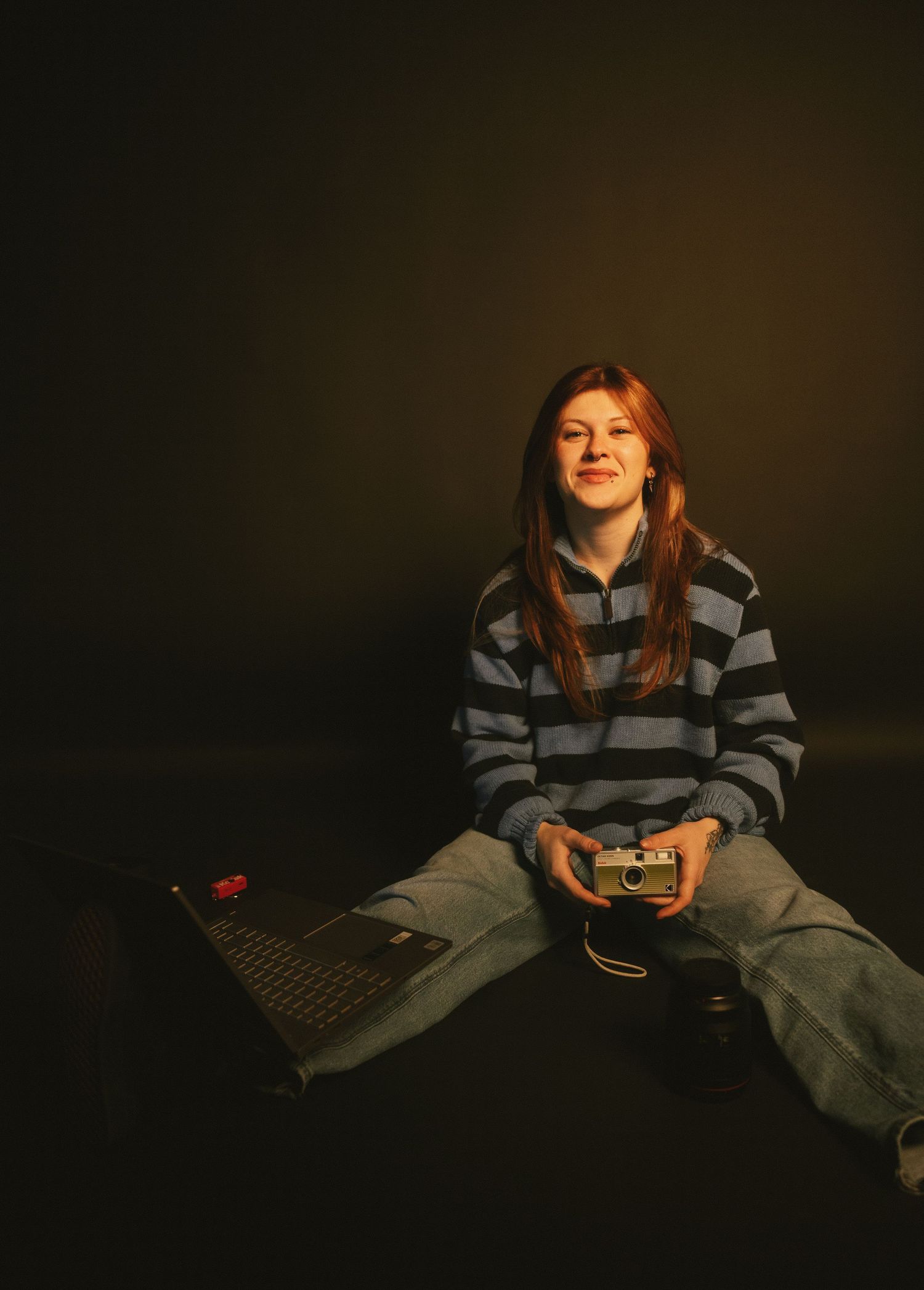 Smiling redhead woman sitting on floor holding vintage camera beside laptop in dark studio.