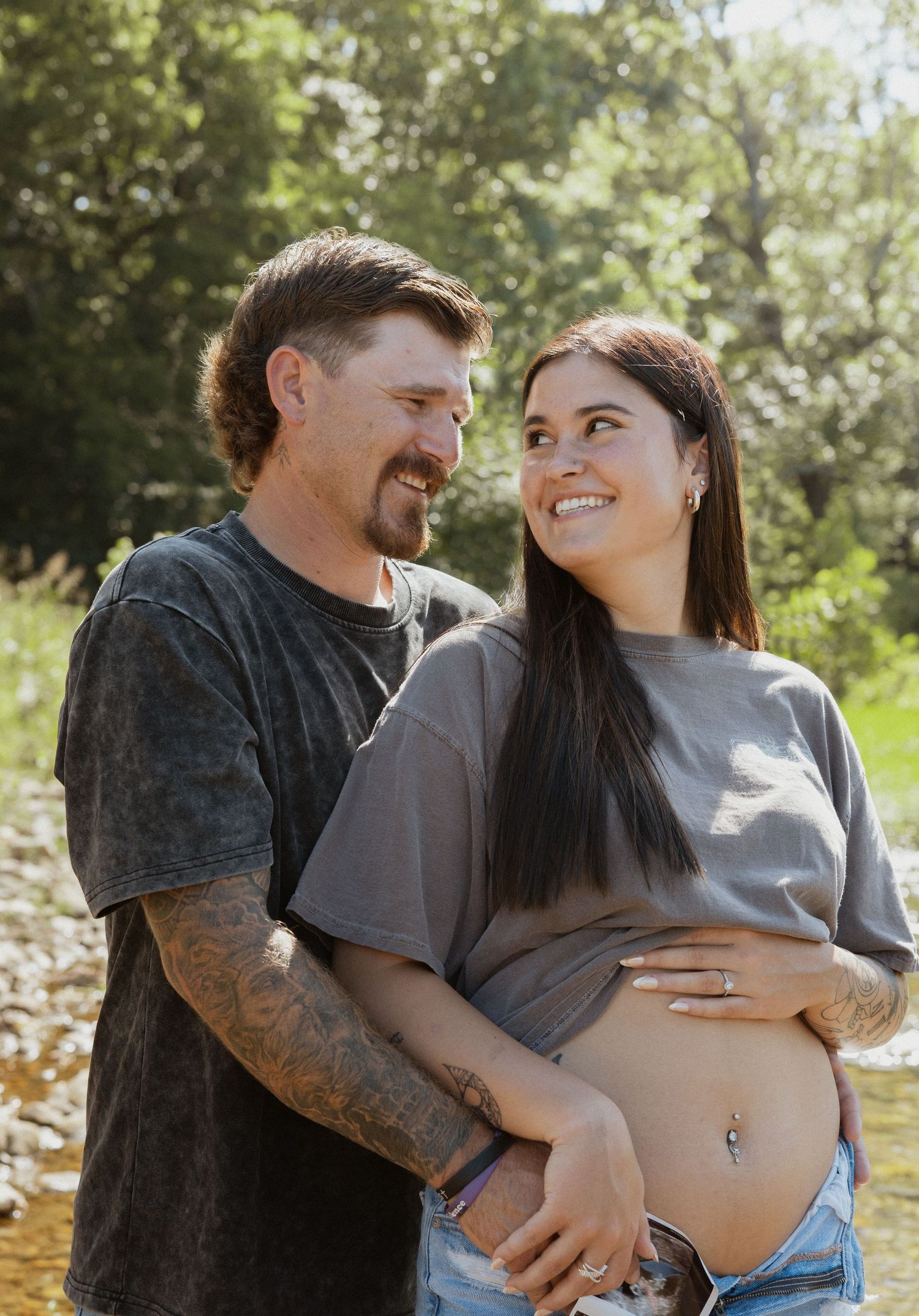 Tattooed couple smiling at each other outdoors, woman showing baby bump near a creek.