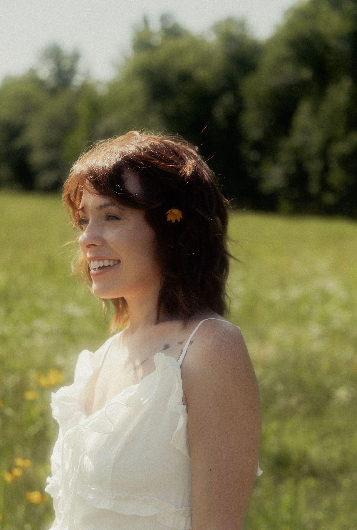 Smiling woman with brown hair and yellow flower accessory wearing white dress in sunny green meadow.