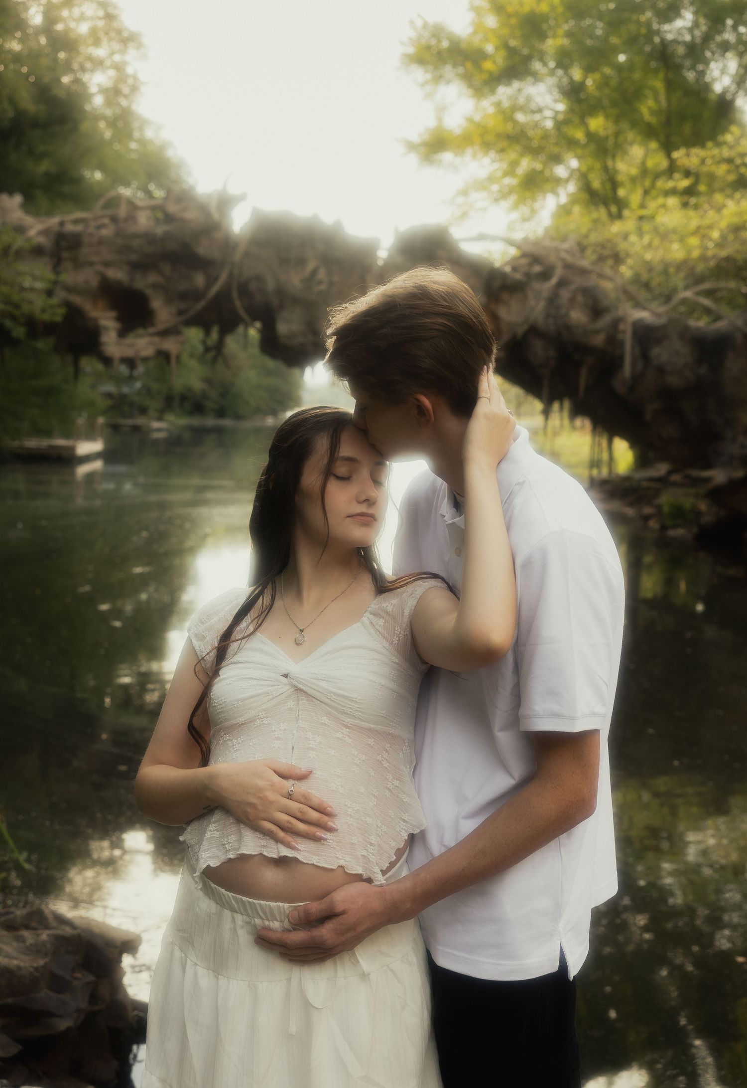 Couple embracing tenderly near a pond, man kissing pregnant woman's head in a dreamy garden.