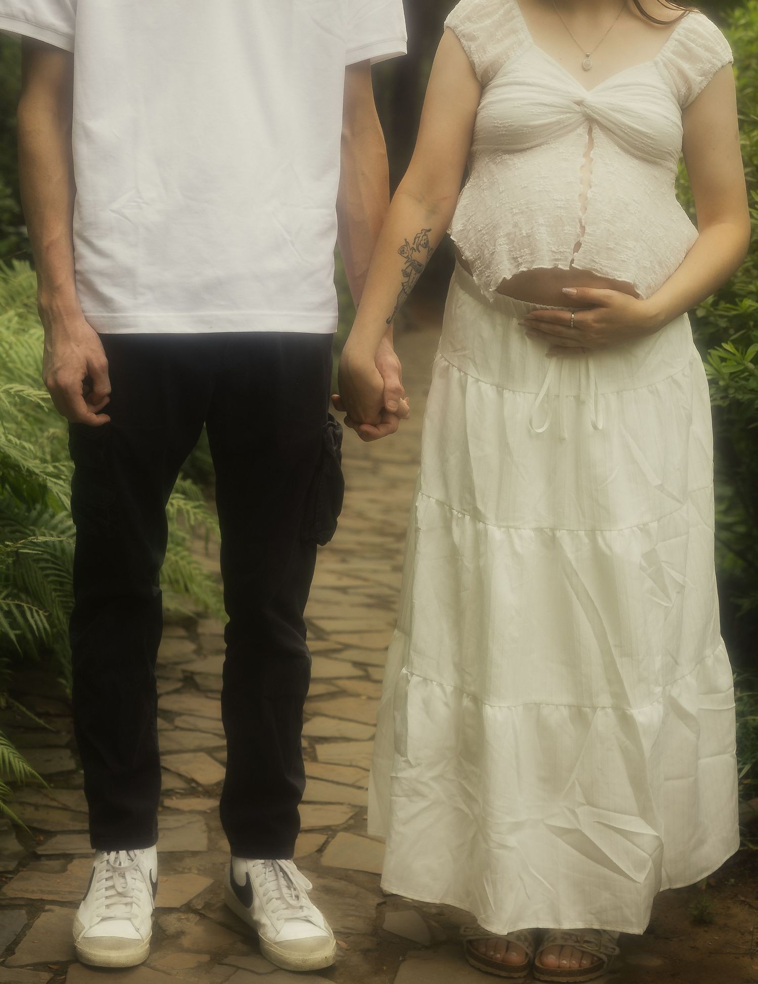Pregnant woman in white dress holding baby bump while holding hands with partner on stone path.