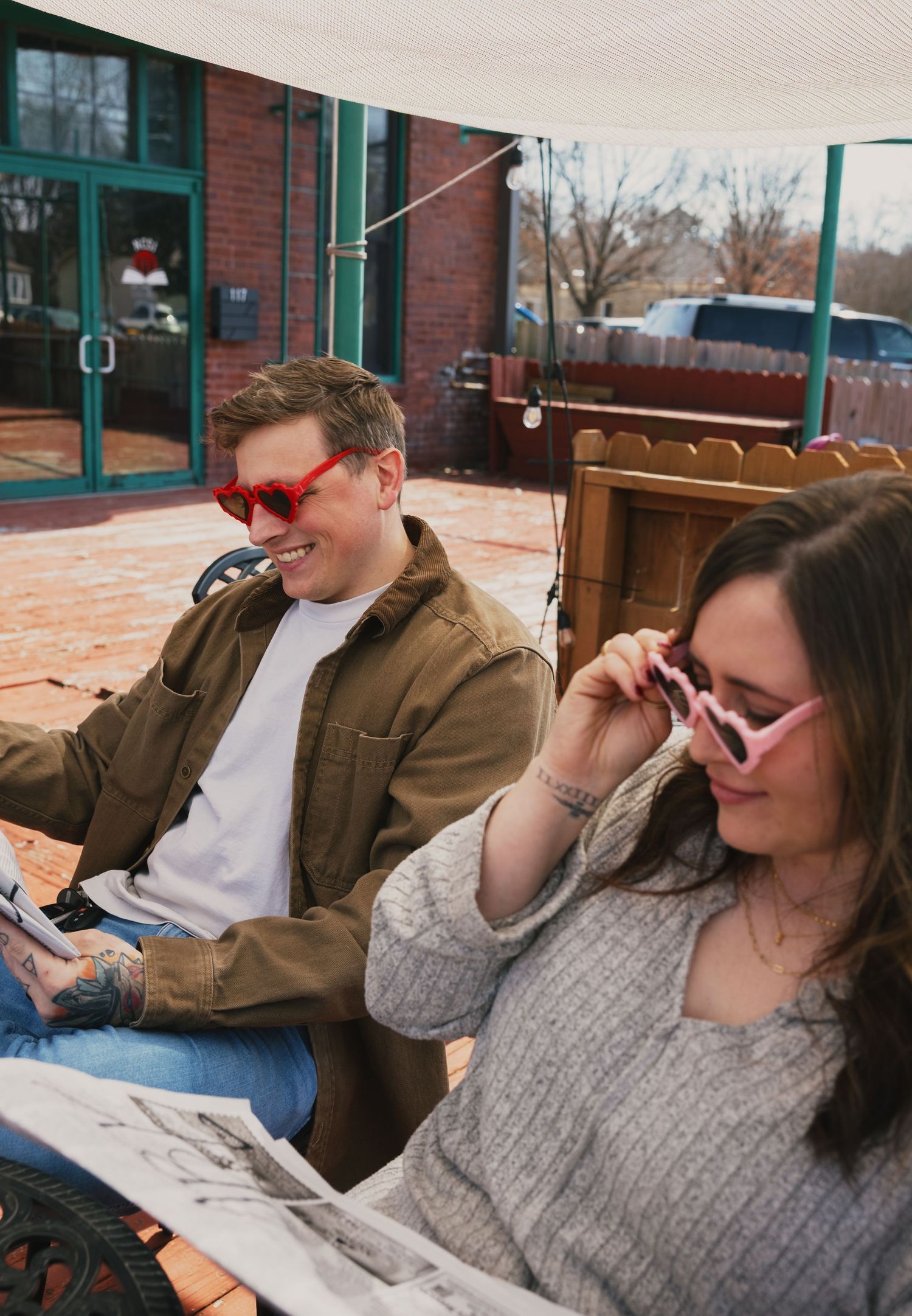Couple wearing colorful heart sunglasses laughing while reading a newspaper on an outdoor patio.