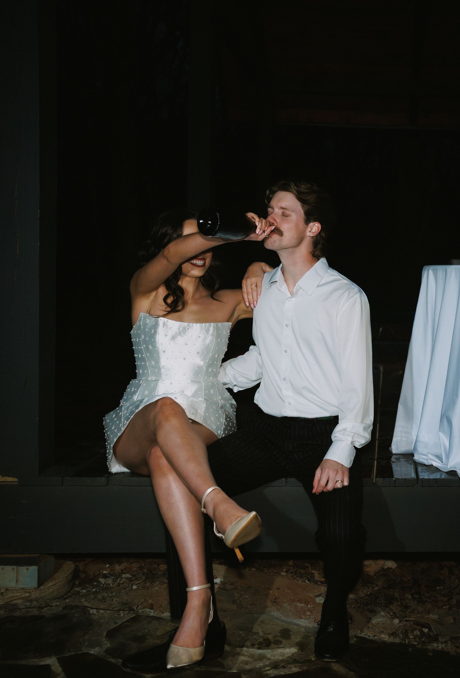 Bride in short white dress and groom in white shirt sharing a drink together at night outdoors.