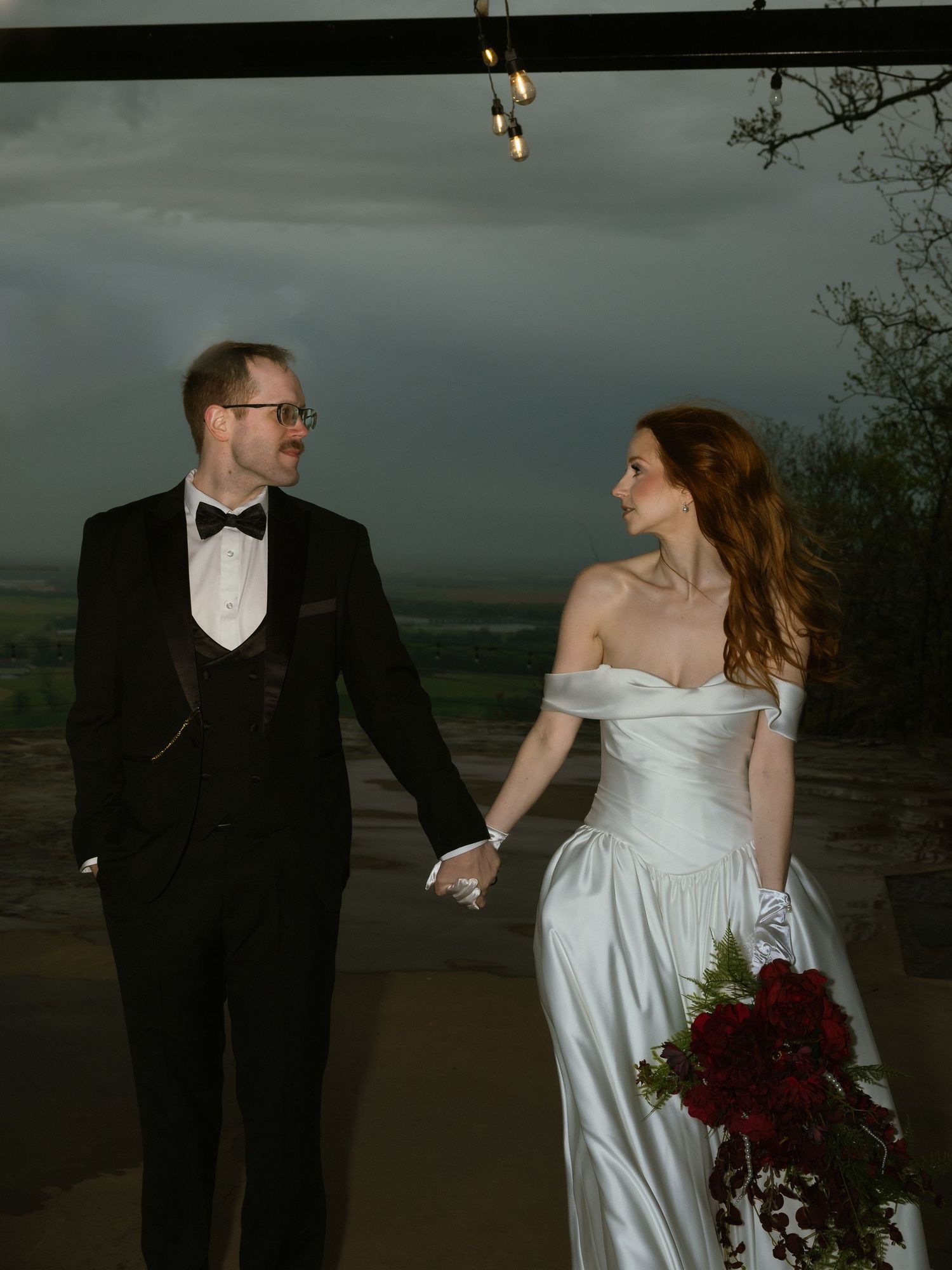 Bride in a satin off-the-shoulder gown holding red roses and a groom in a black tuxedo hold hands under a stormy sky at an outdoor wedding.