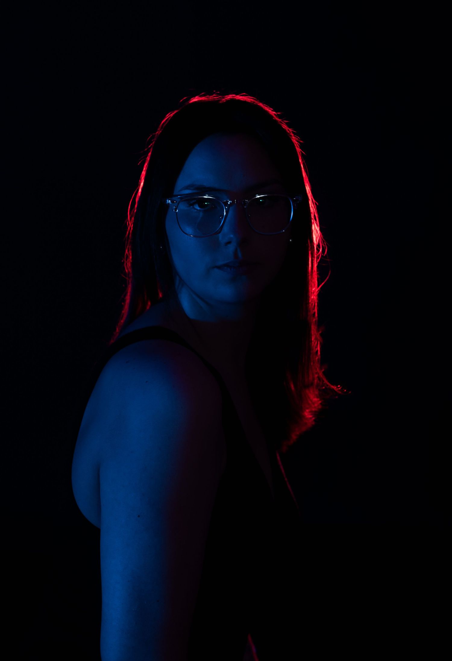 Dramatic portrait of woman with glasses illuminated by red and blue neon lights on dark background.