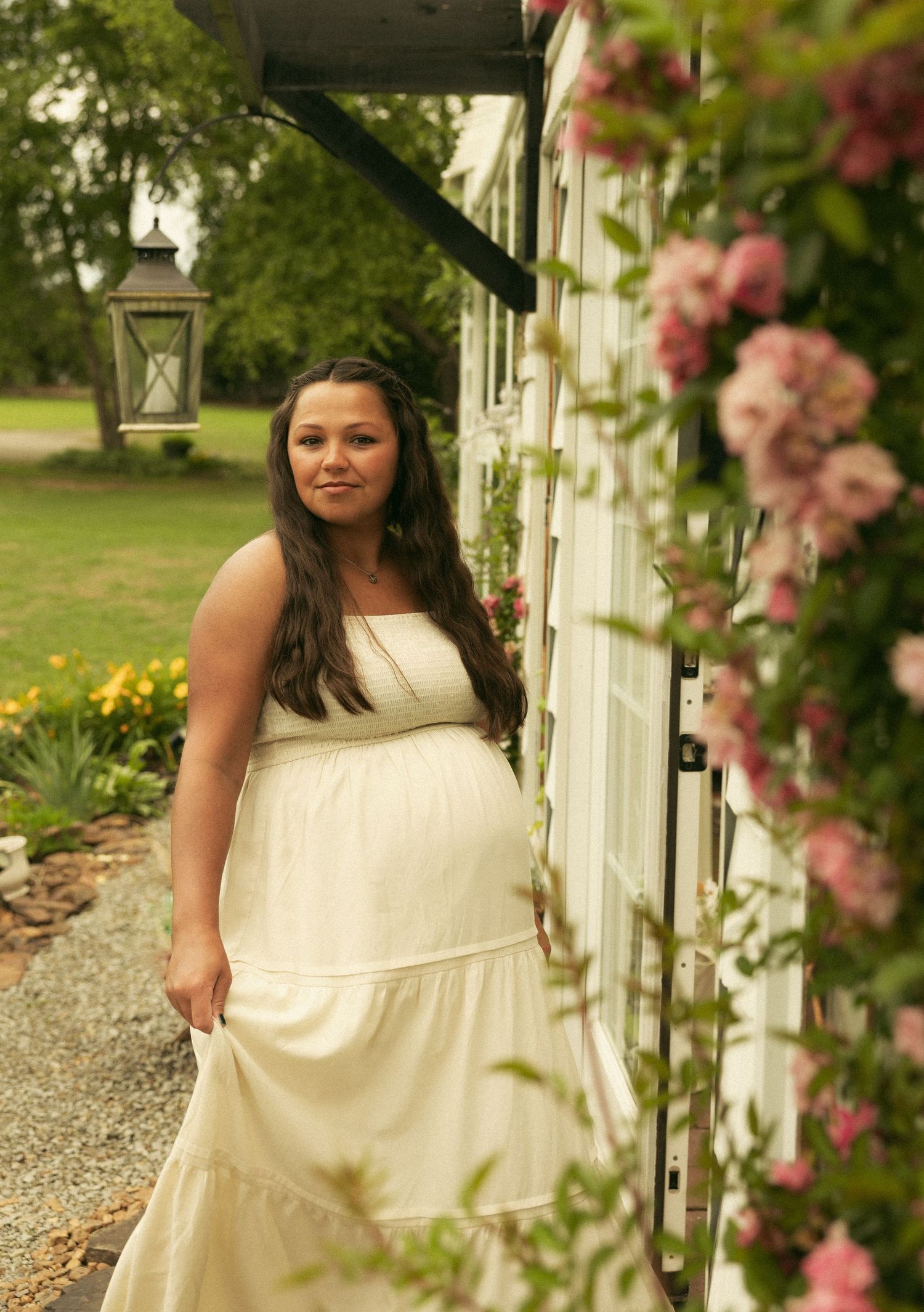 Pregnant woman in flowing white dress standing by roses near a white building with lanterns in a garden.