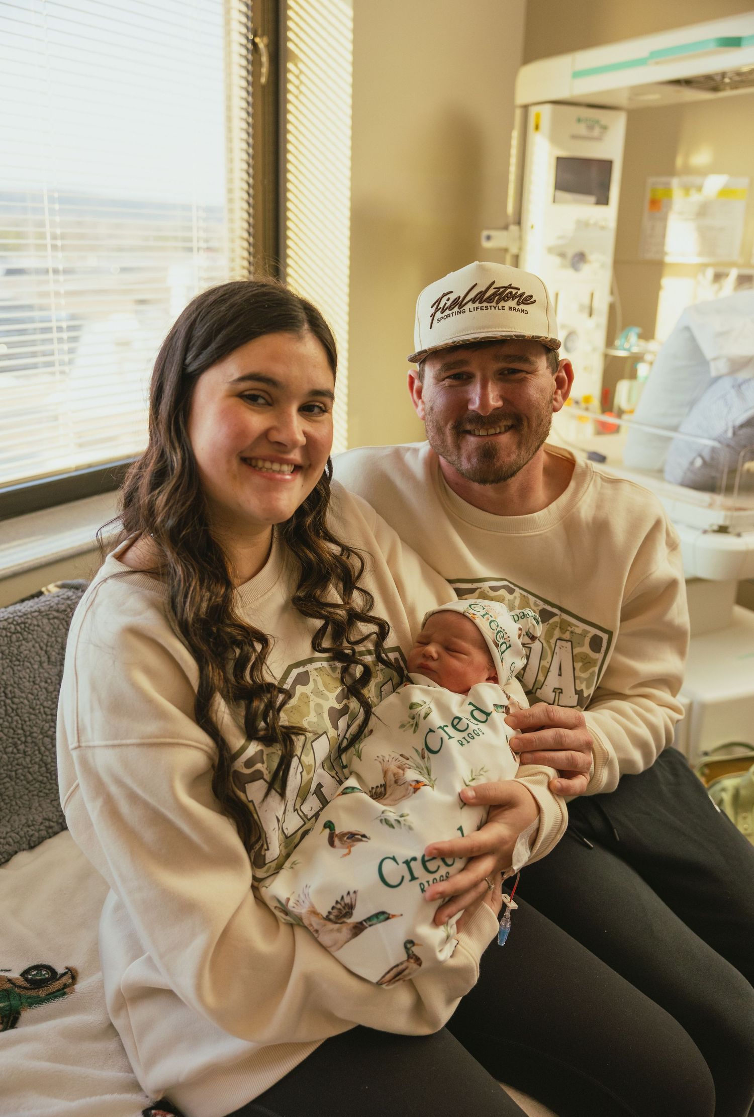 Happy new parents pose with their swaddled newborn baby in a hospital room, both wearing matching cream hoodies.