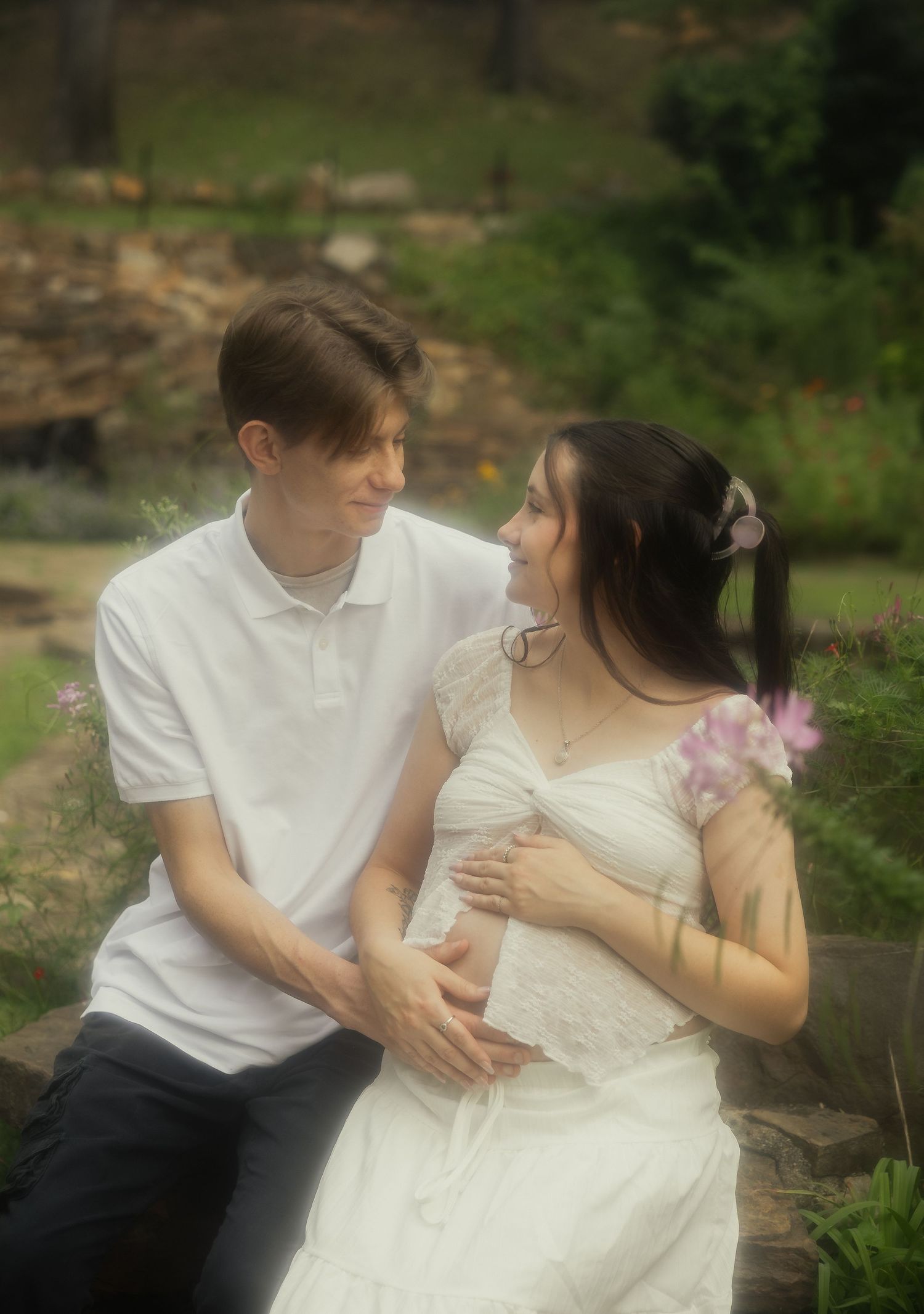 Couple sitting together outdoors, cradling pregnant belly surrounded by wildflowers and greenery.