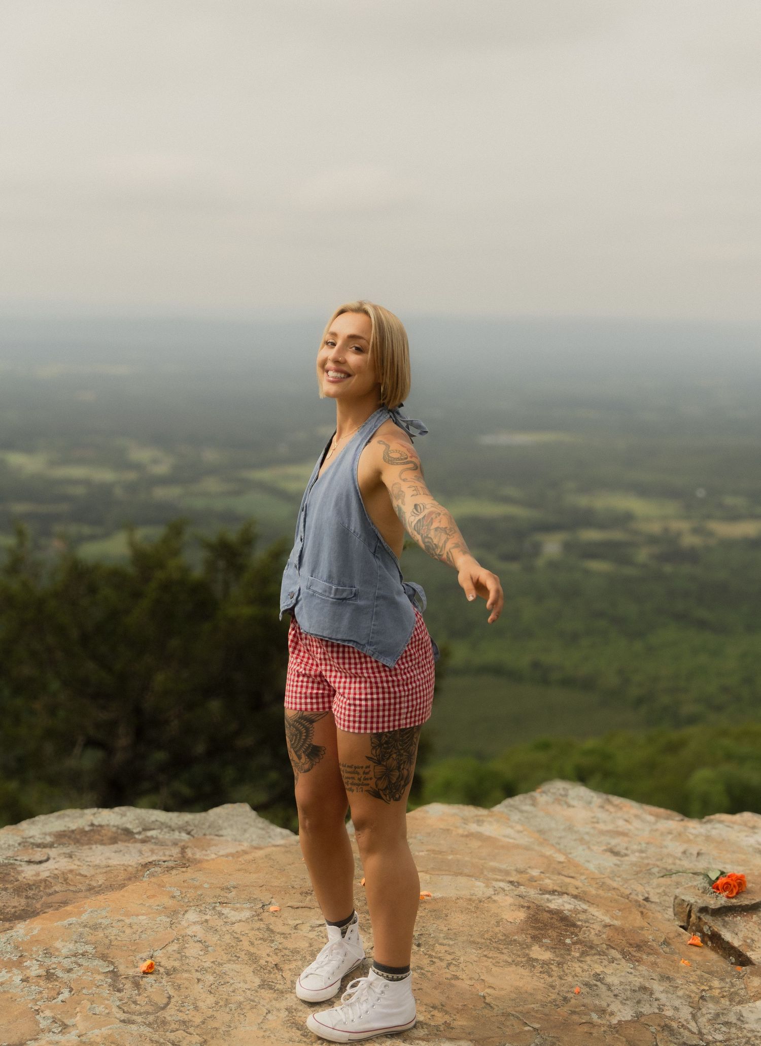 Tattooed blonde woman in denim vest and plaid shorts posing on mountain overlook with scenic valley view.