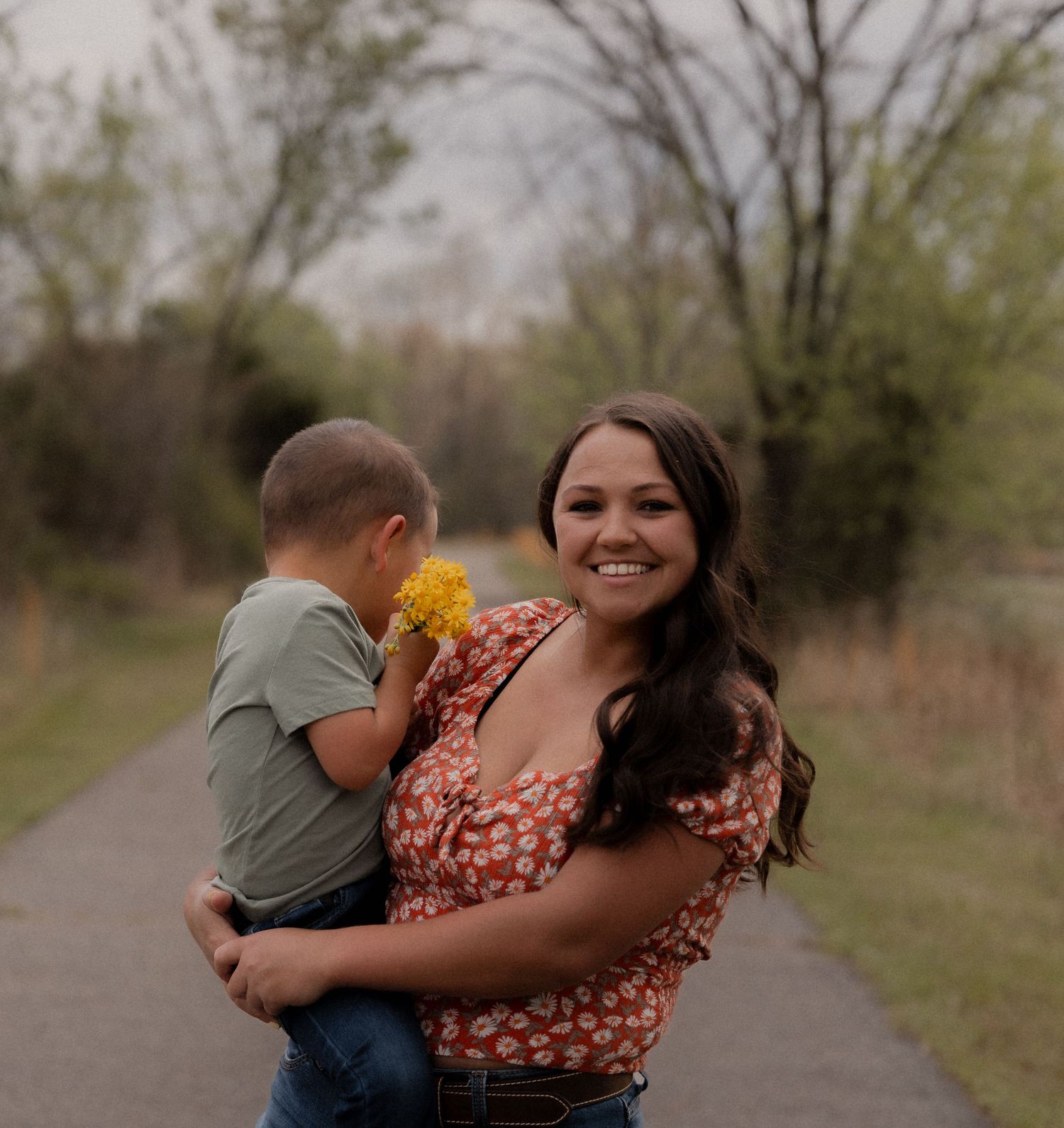 Smiling woman holding a young boy with yellow flowers on a tree-lined path.