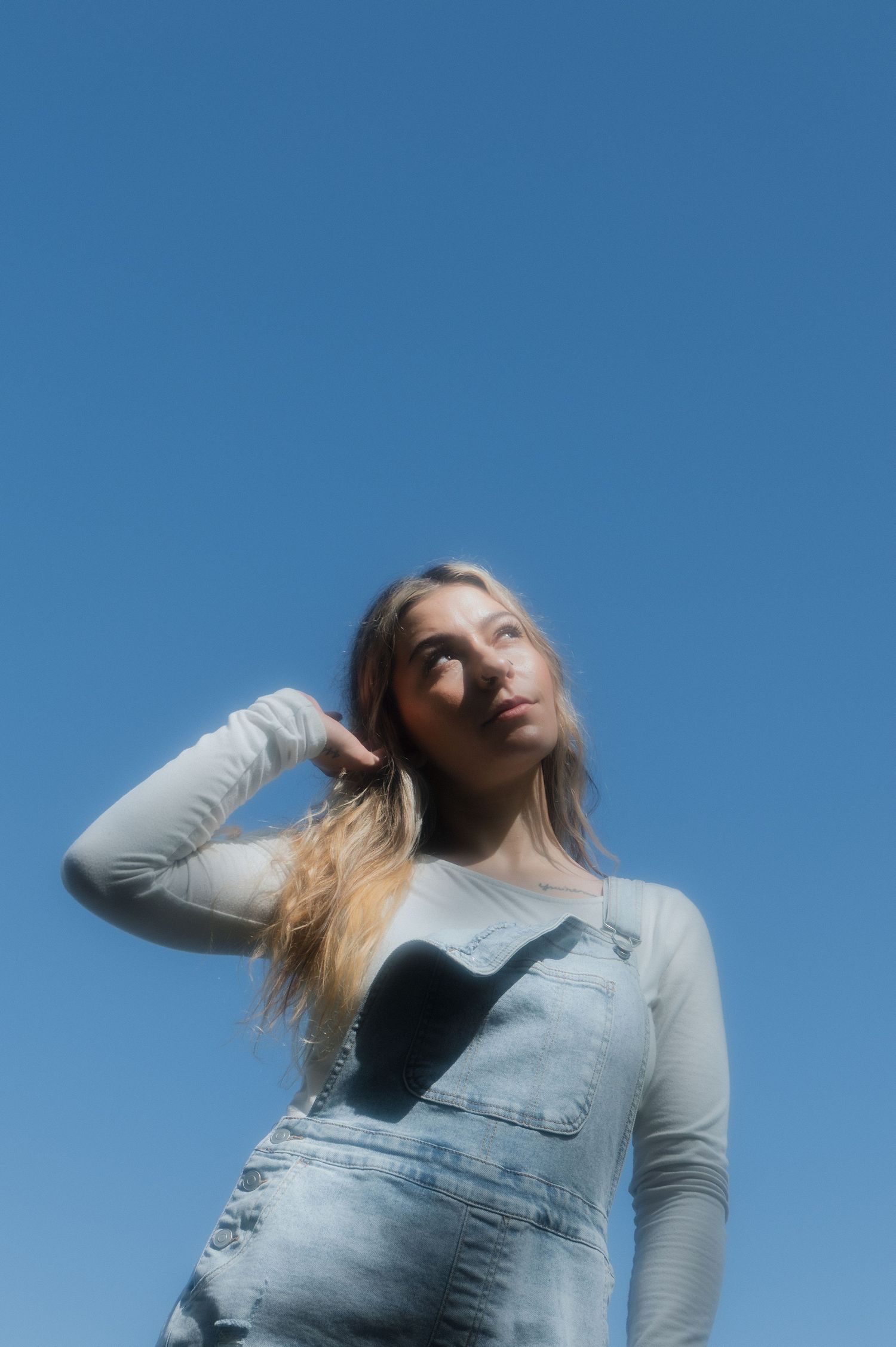 Blonde woman in denim overalls holding hair back against clear blue sky background.