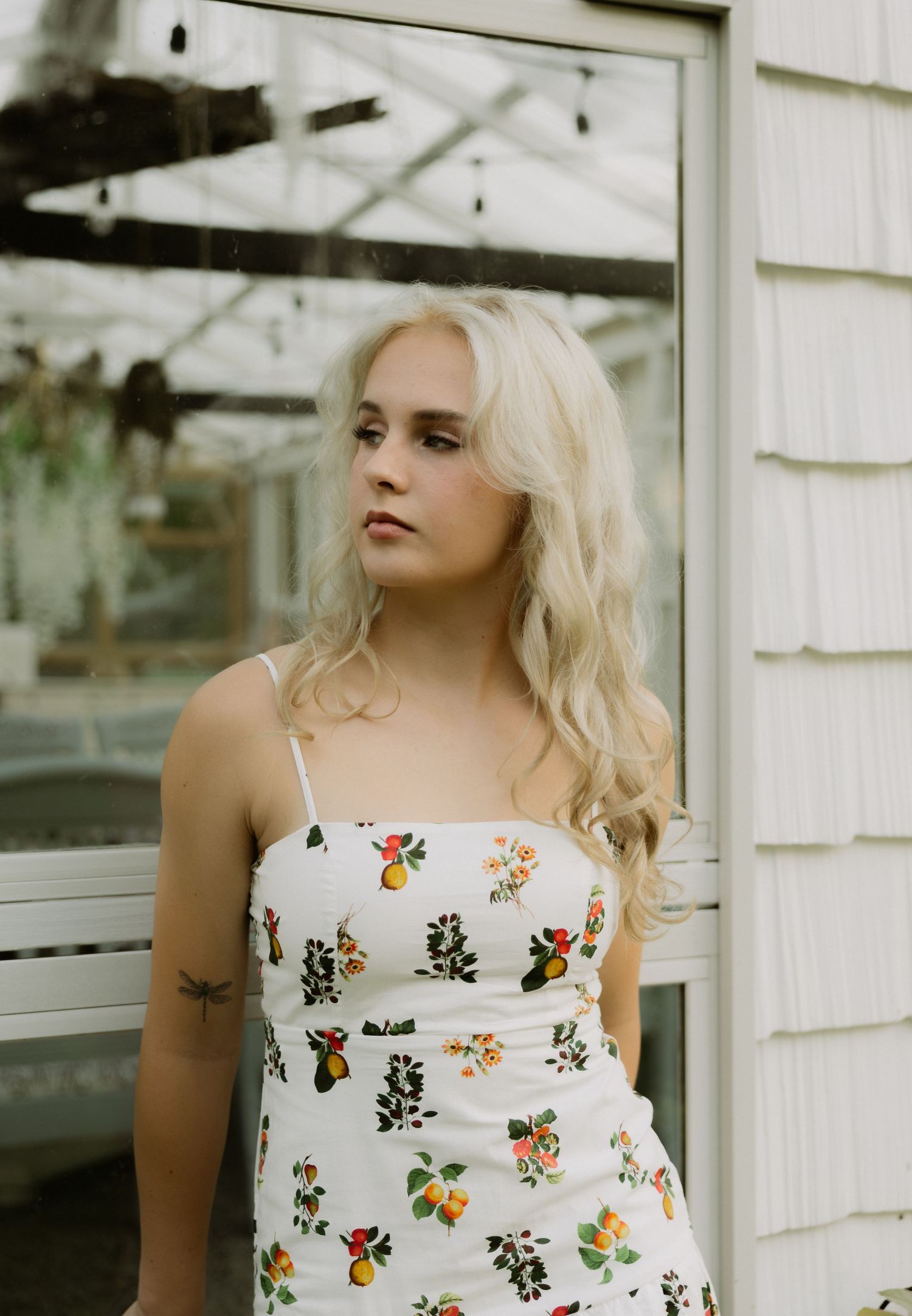 Young blonde woman in floral sundress standing near a greenhouse window outdoors.