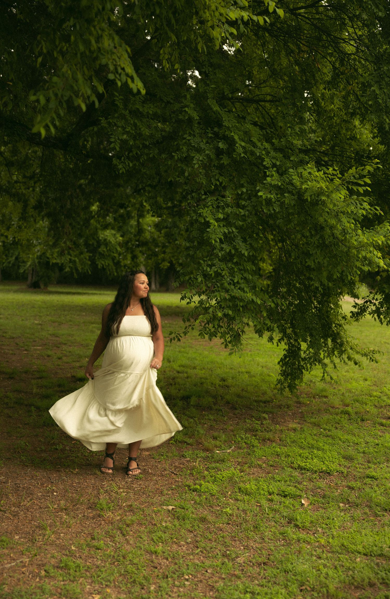 Pregnant woman in flowing white dress standing under large green tree.