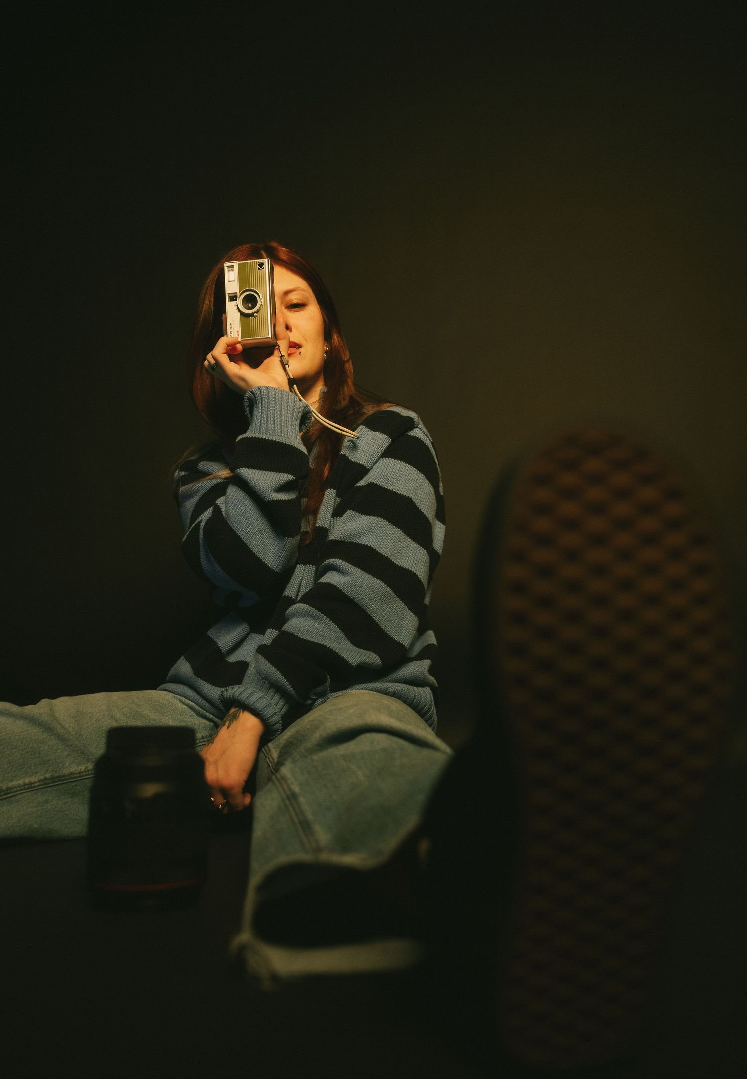 A young woman in a striped sweater holds a vintage camera up to her face in a moody, dimly lit studio.