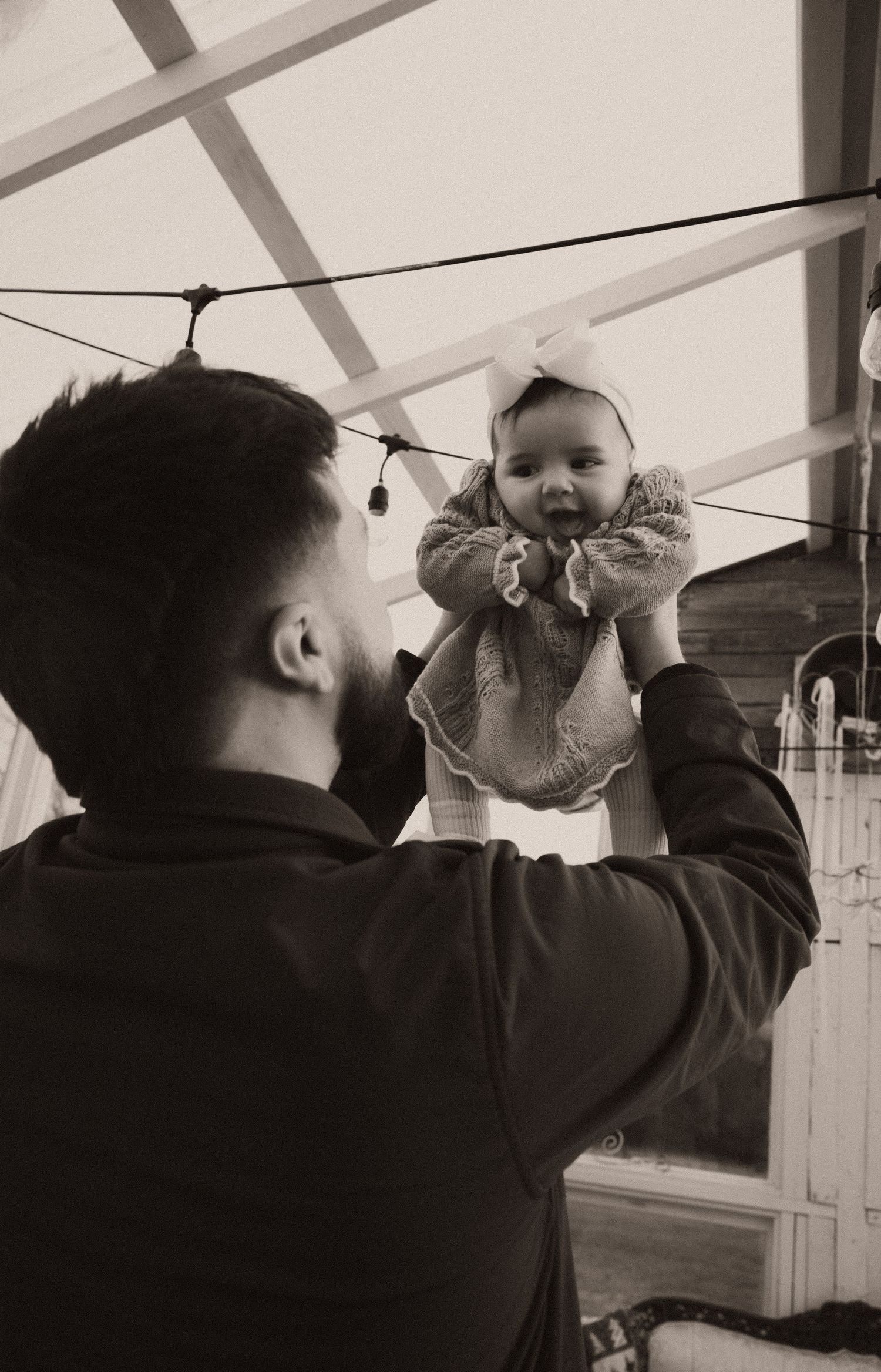 Black and white photo of father lifting smiling baby girl with bow headband in greenhouse.