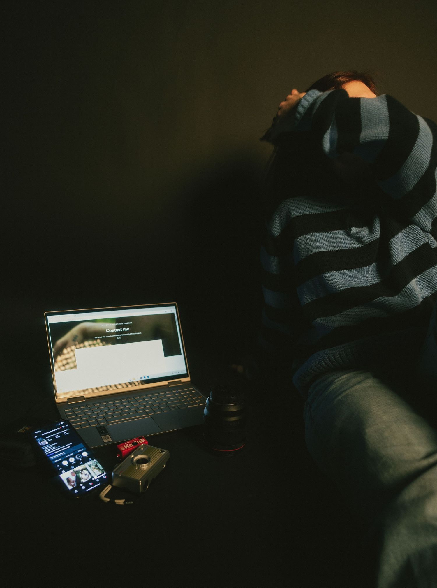 Person in striped sweater sitting in dark room with laptop and camera gear.