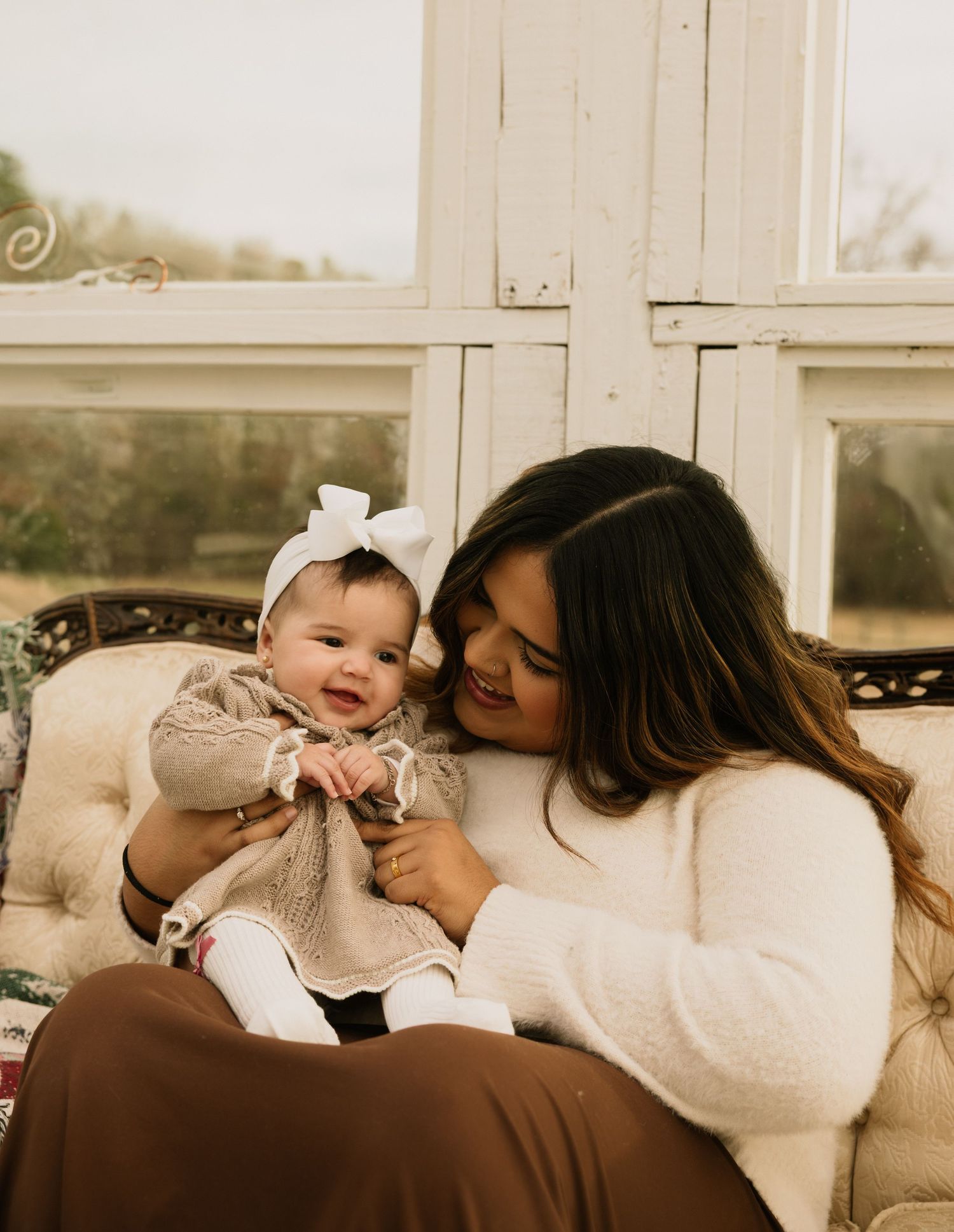 A joyful mother sits on a vintage sofa smiling at her happy baby girl wearing a white bow headband.