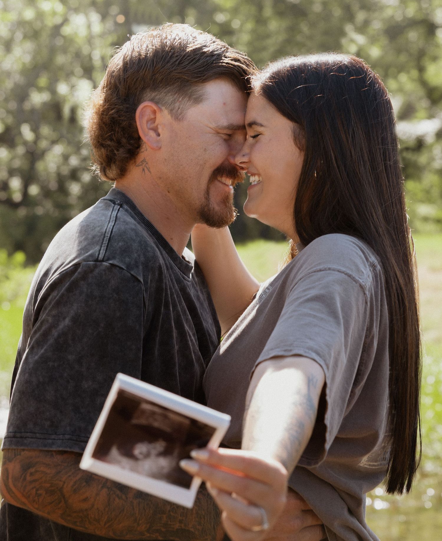 Happy couple nearly kissing outdoors while holding an ultrasound photo in sunny natural light.