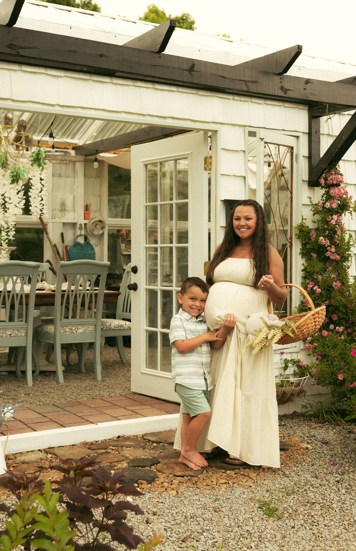 Smiling pregnant woman in white dress holding basket with young boy outside a charming white greenhouse.