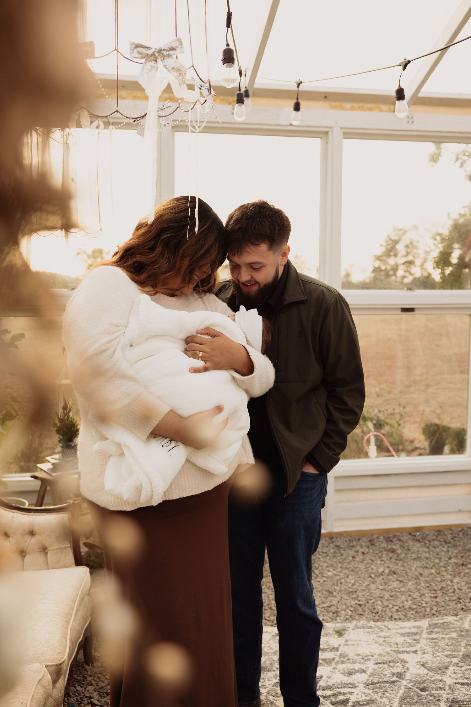 A couple shares a tender moment with their newborn baby in a warmly lit greenhouse setting.