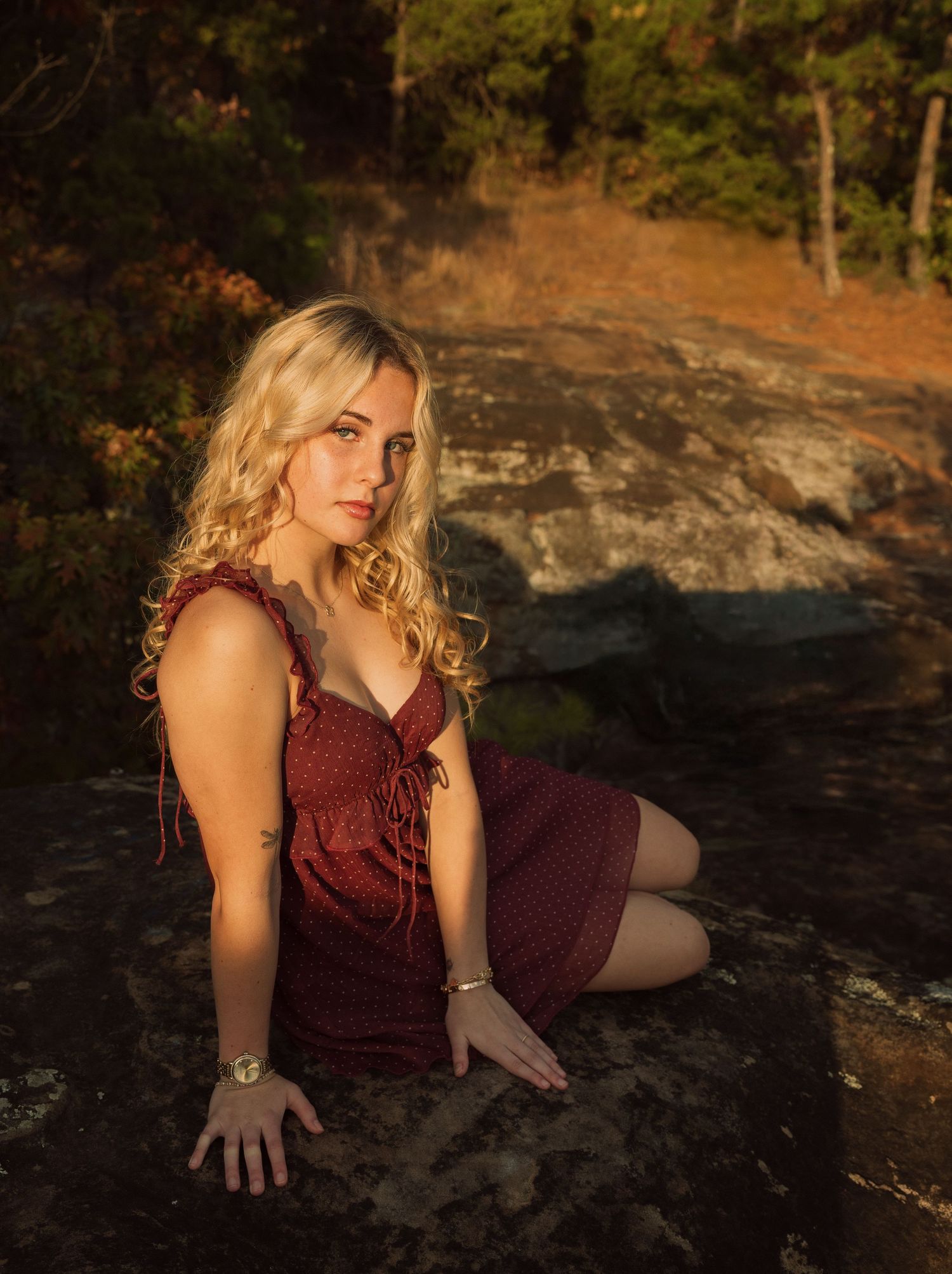 Young blonde woman in red dress sitting on rock during golden hour in autumn forest setting.