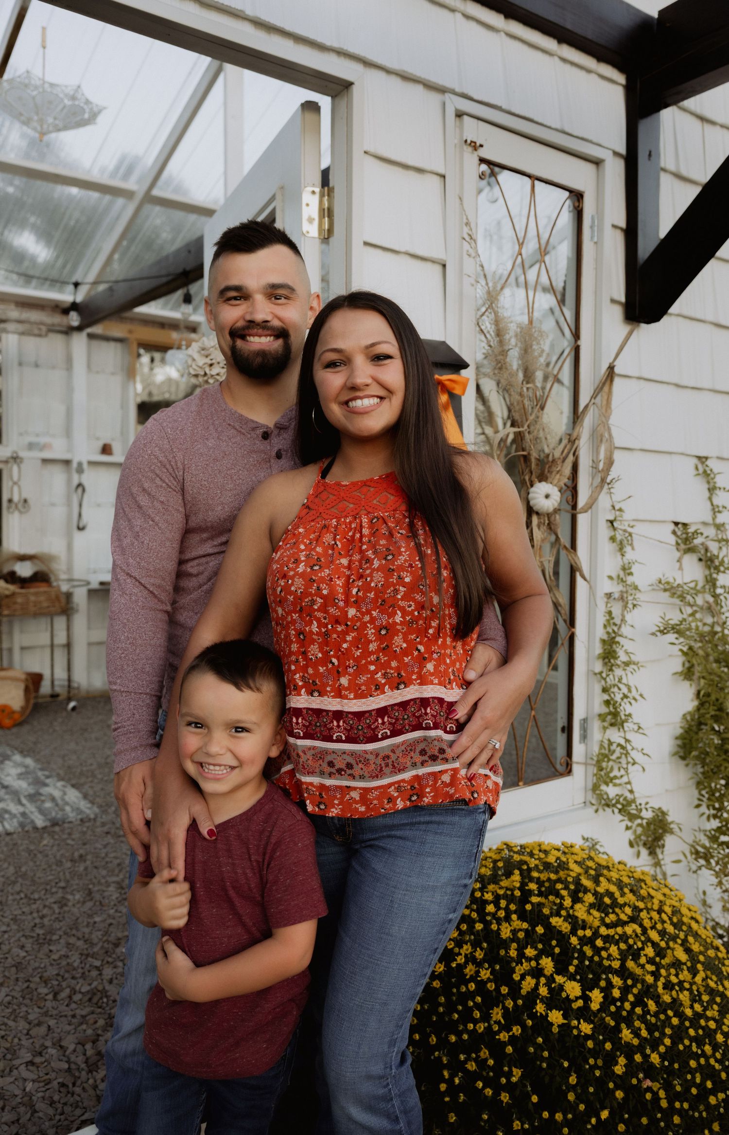 A smiling family of three poses outside a charming white building surrounded by fall flowers and autumn decor.