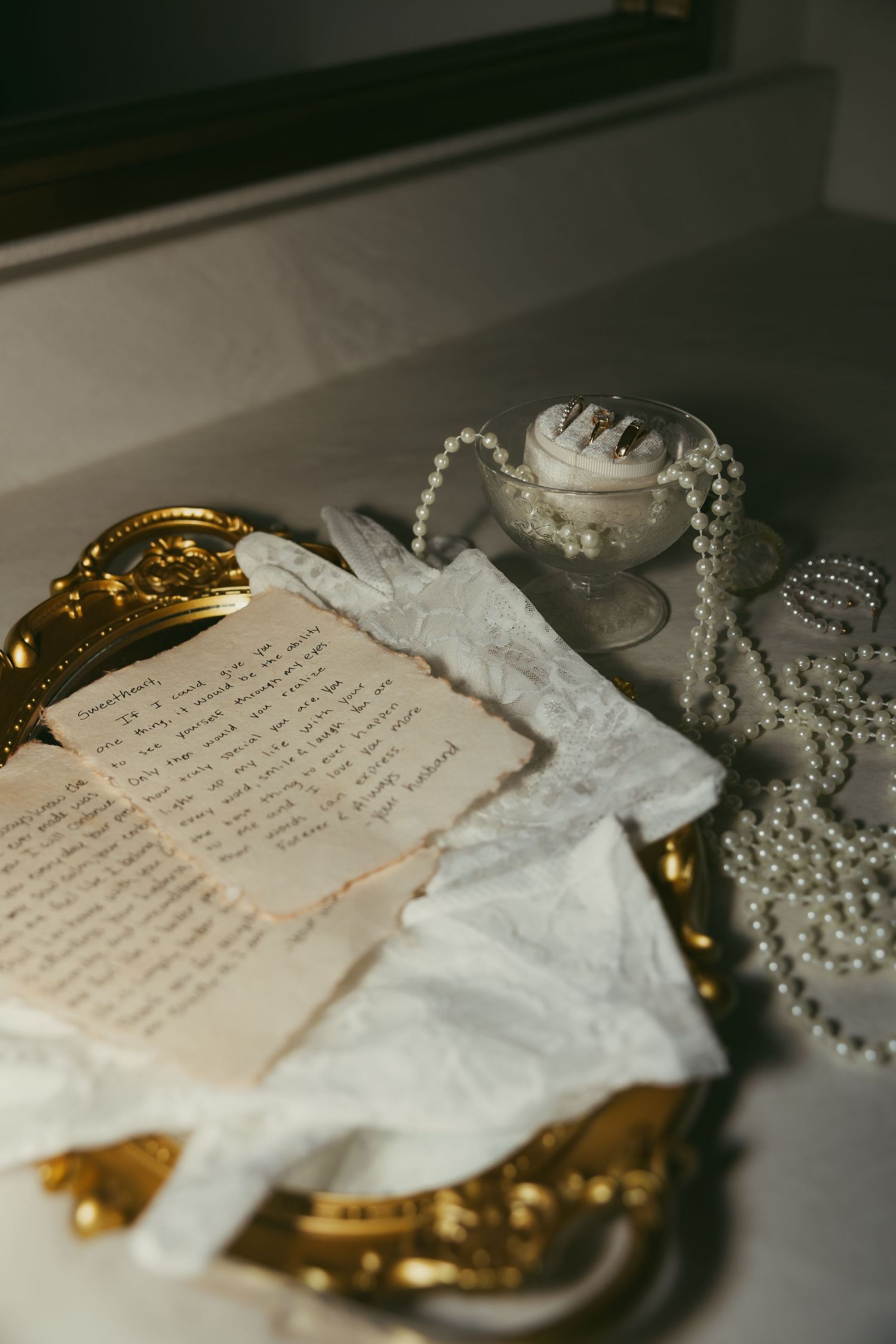 Handwritten love letter on gold tray with white lace gloves, pearl necklace, and rings on marble surface.