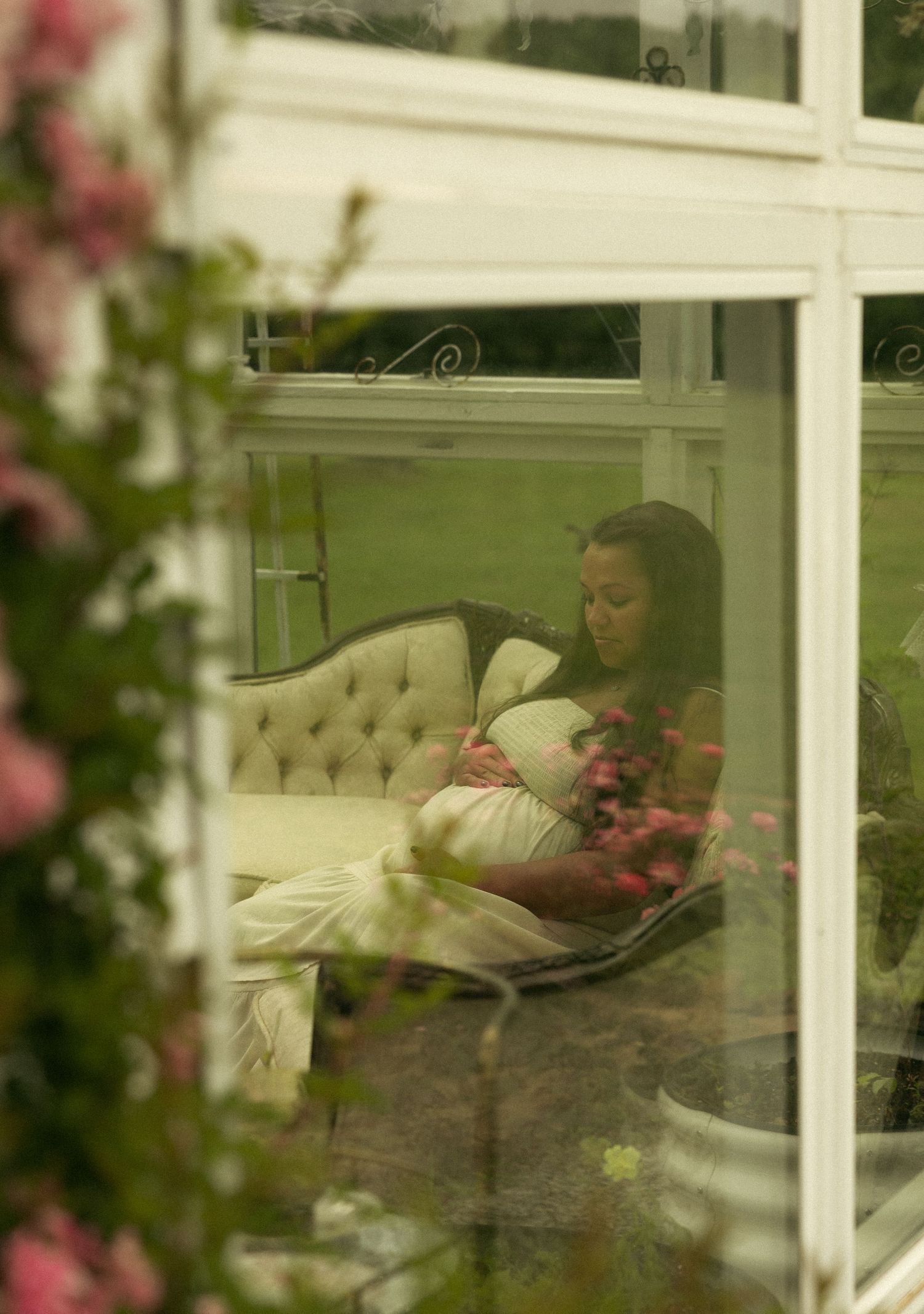 Woman in white outfit cradling bump on vintage sofa viewed through greenhouse window with pink flowers.