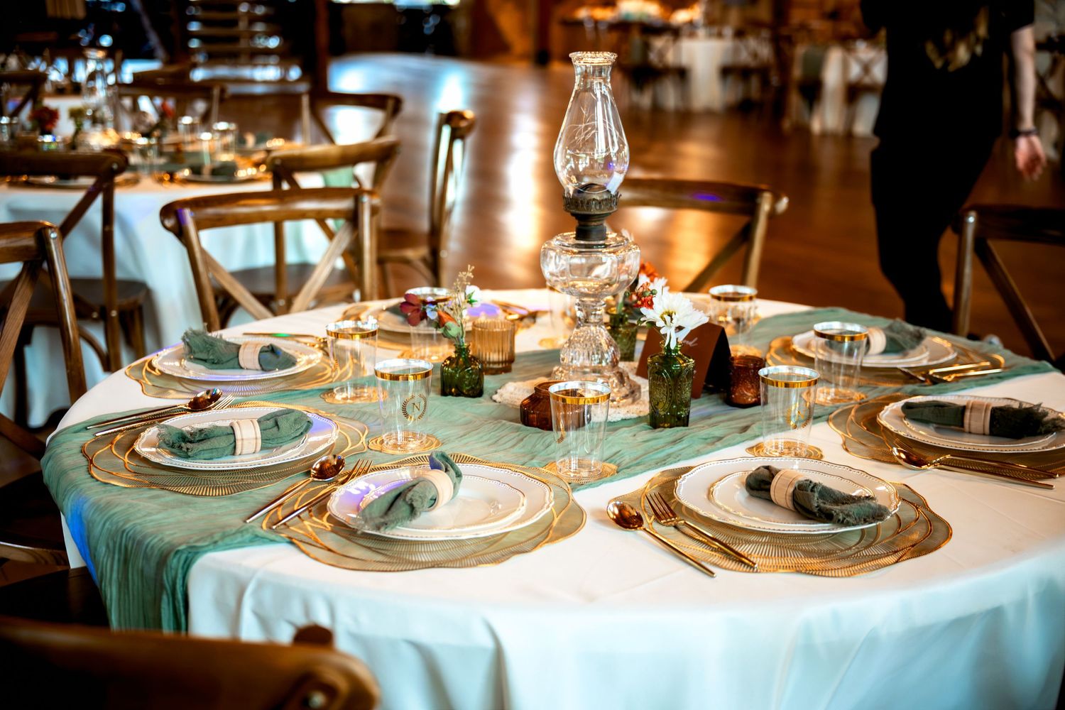 Elegant wedding reception table with teal linen, gold chargers, folded napkins, and a vintage oil lamp centerpiece.