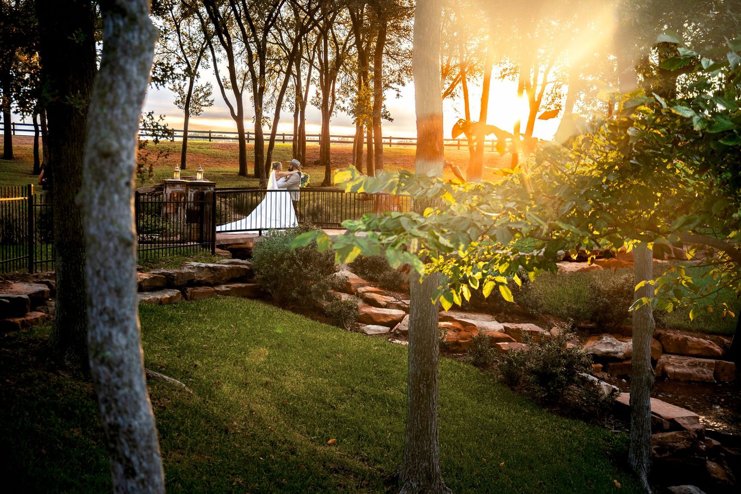 Golden sunset illuminates a lush garden with stone steps, trees, and a couple in the distance at an outdoor wedding venue.