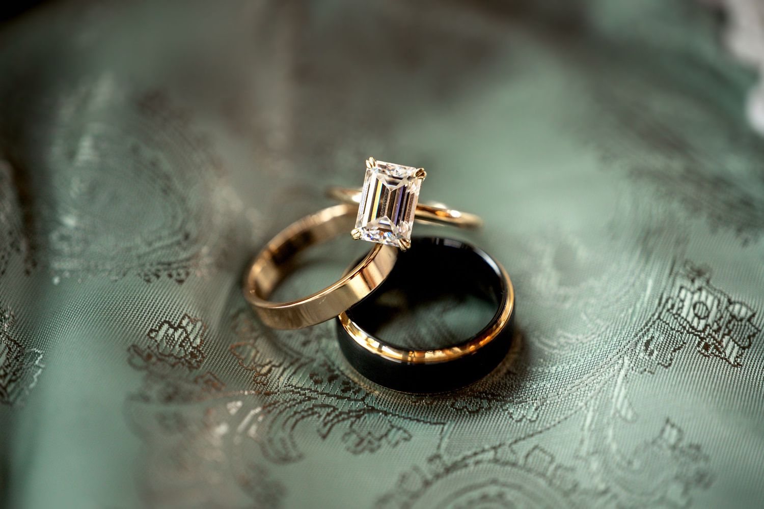 Gold engagement ring with emerald-cut diamond solitaire resting on a black wedding band, displayed on green fabric.