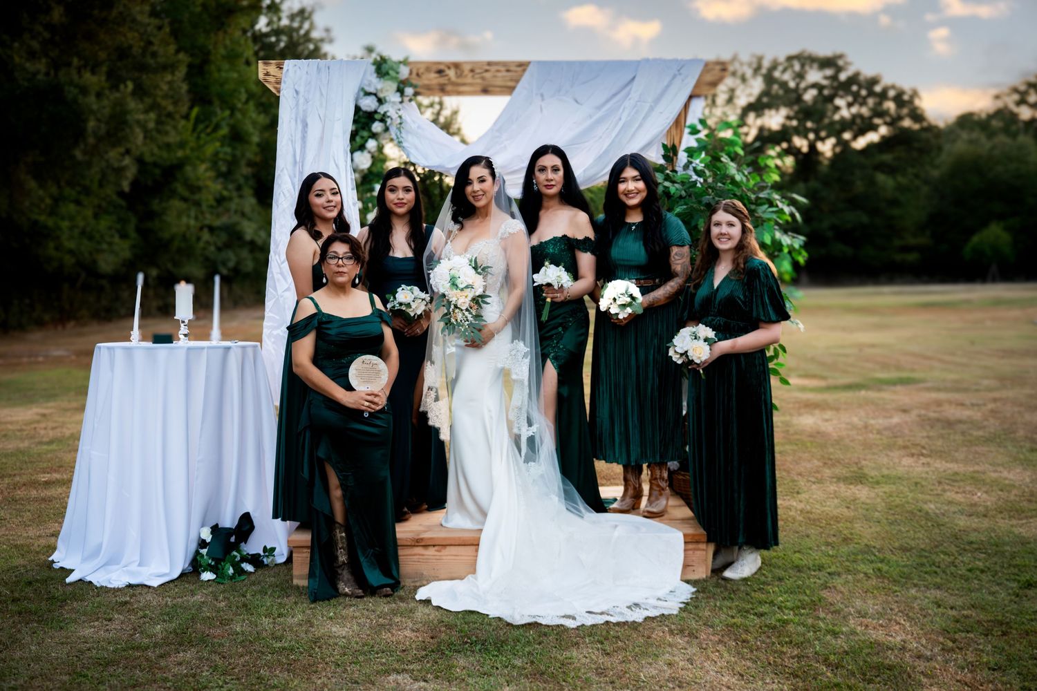Bride posing with bridesmaids in emerald dresses at outdoor wedding ceremony.