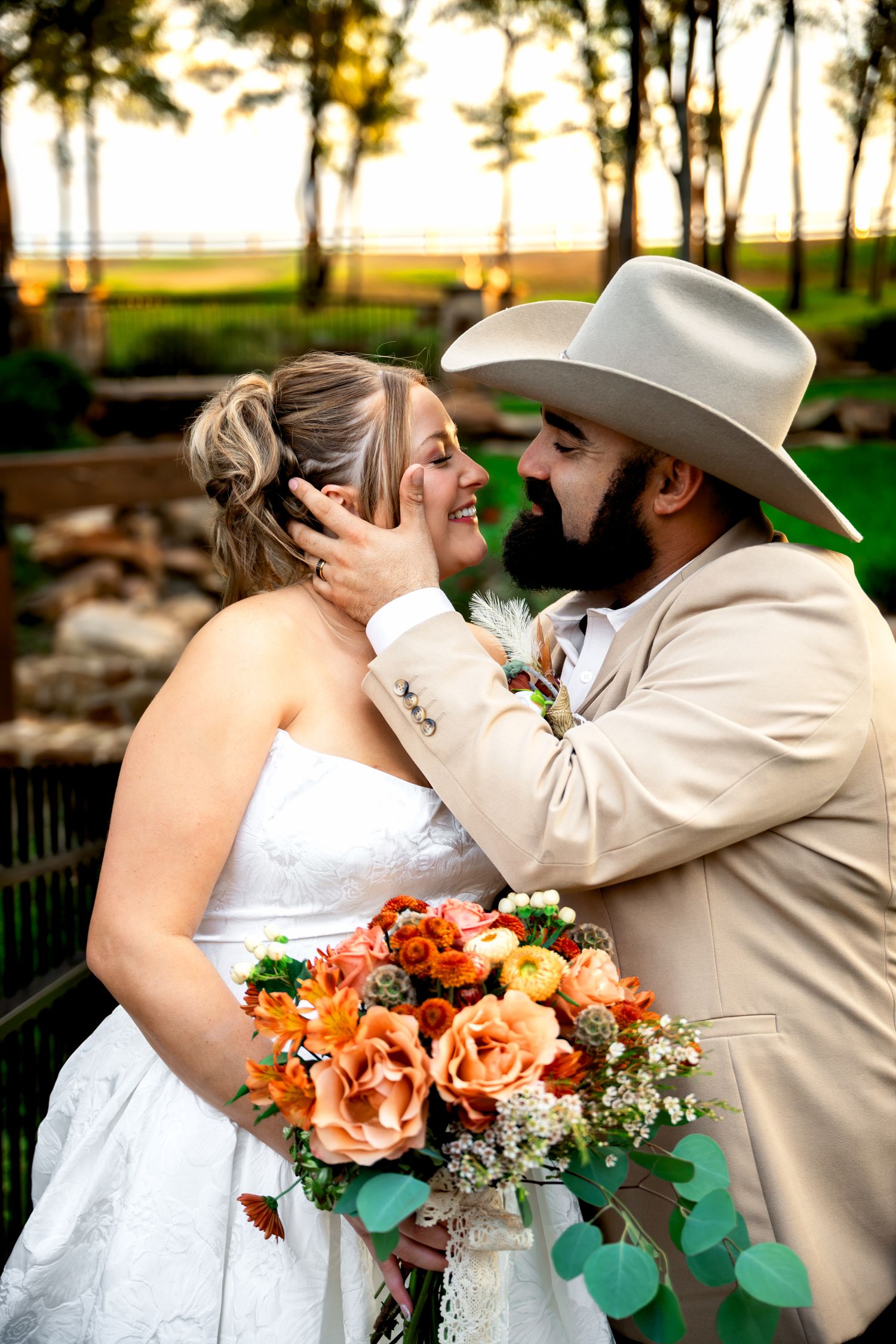 A groom in a tan cowboy hat tenderly cups his bride's face as she holds an orange floral bouquet at their outdoor wedding.