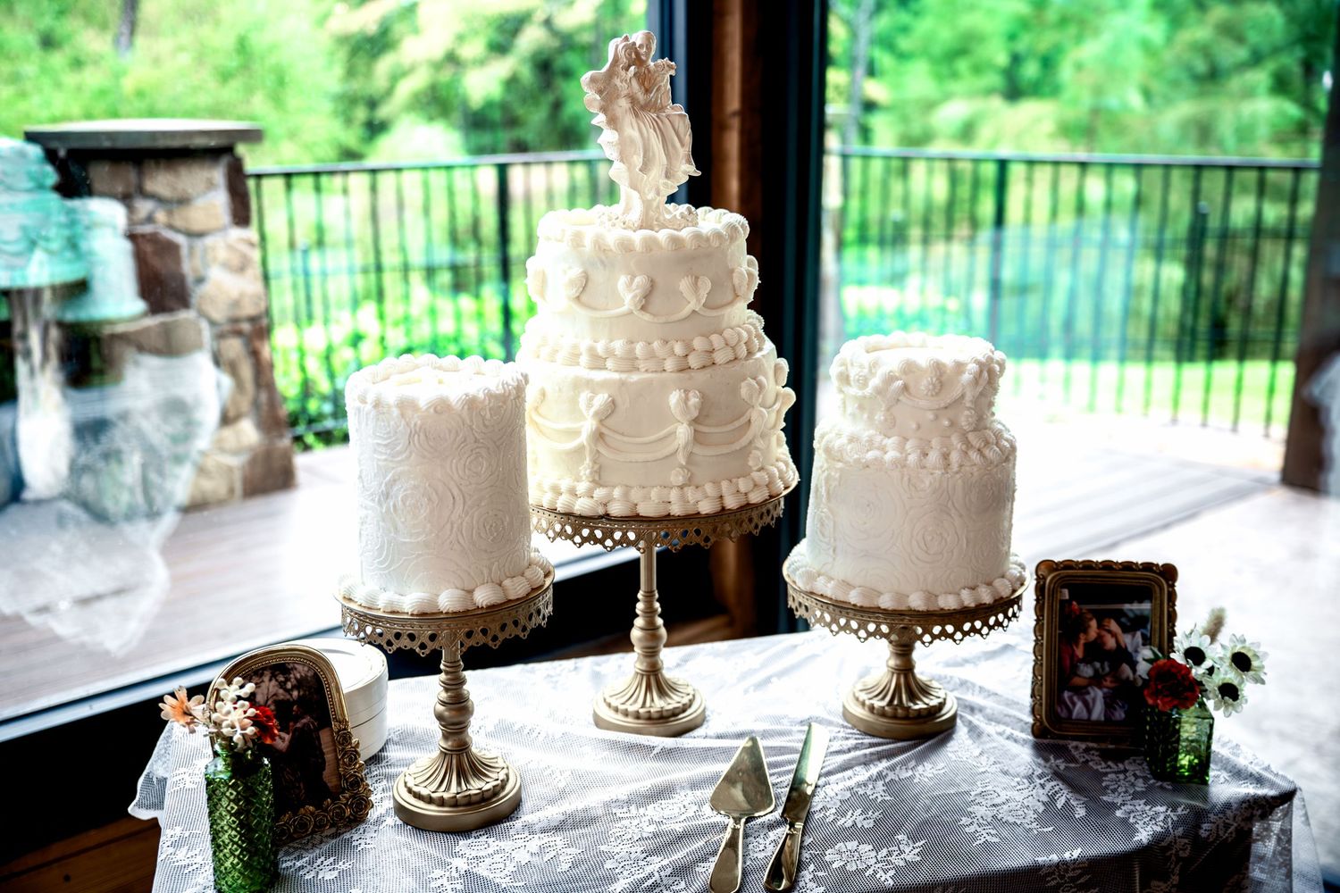 Three white wedding cakes on gold stands displayed on a table with lush greenery visible through windows behind them.