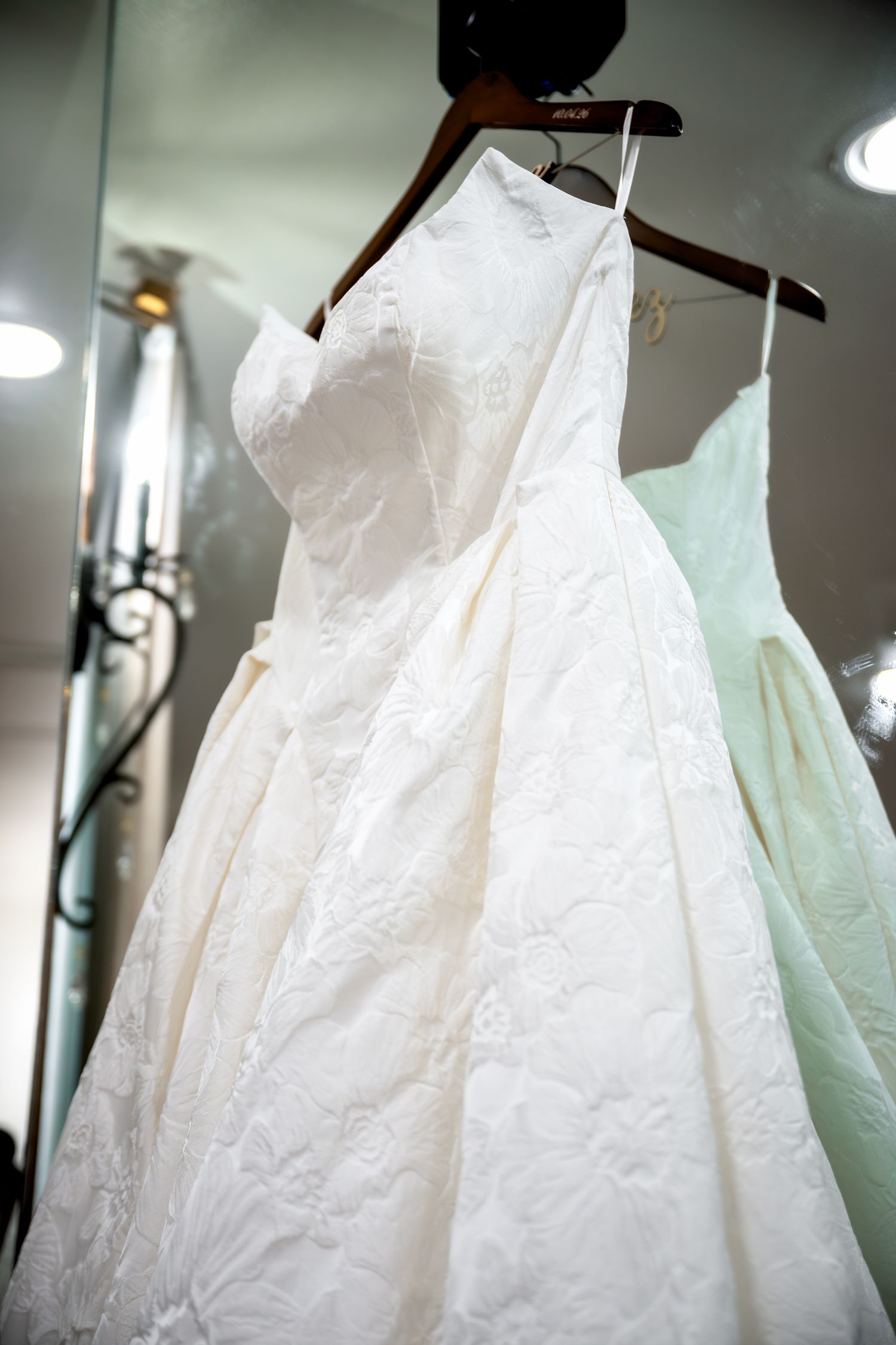 White lace ball gown wedding dress hanging on wooden hanger in bridal boutique with mint green dress in background.