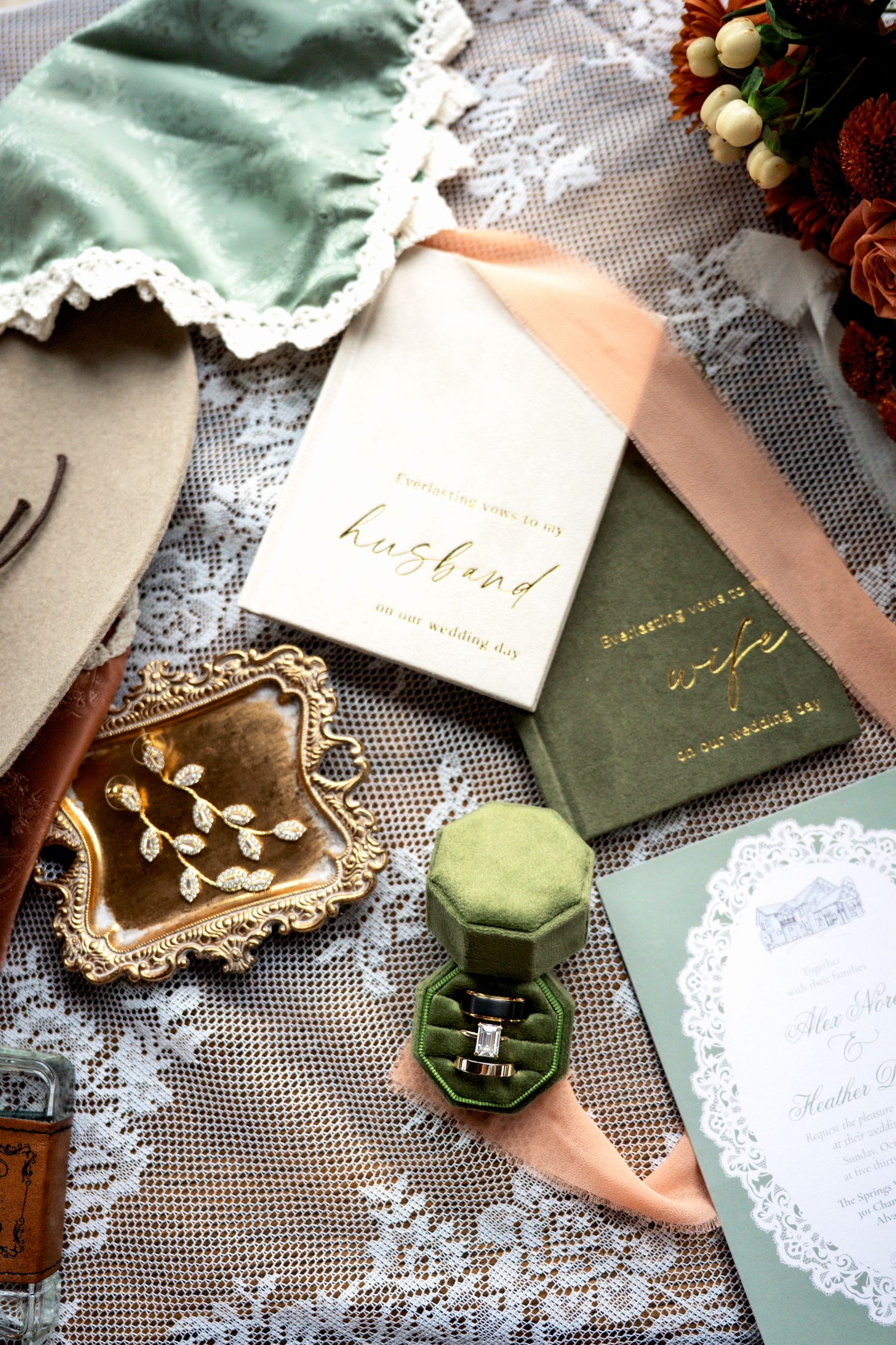 Elegant wedding flat lay with green velvet ring box, gold jewelry tray, vow books, lace, and floral accents.