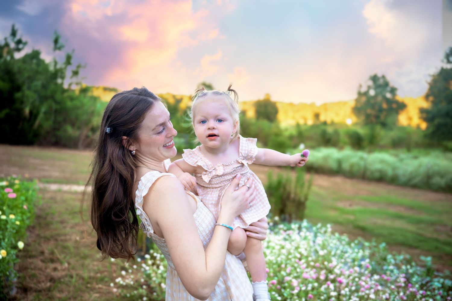 Mother holds baby girl in pink dress in a flower field at sunset.