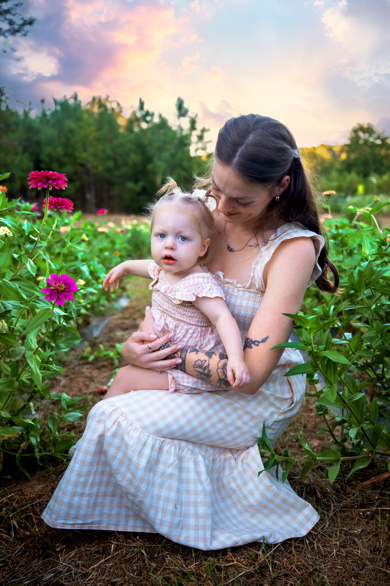 Young mother holding baby girl among colorful zinnia flowers at sunset.