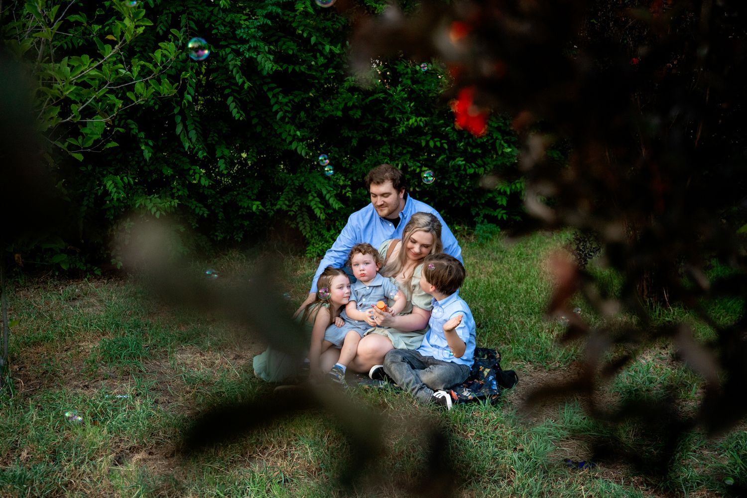 Happy family of five sitting together on grass surrounded by lush garden greenery.