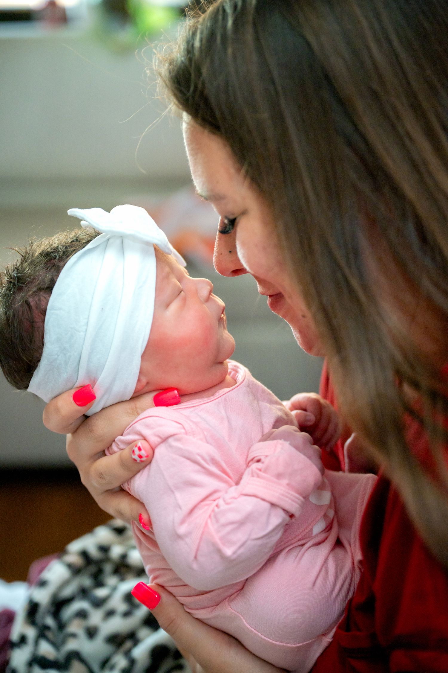 New mother with red nails tenderly holding newborn baby girl in white bow headband.