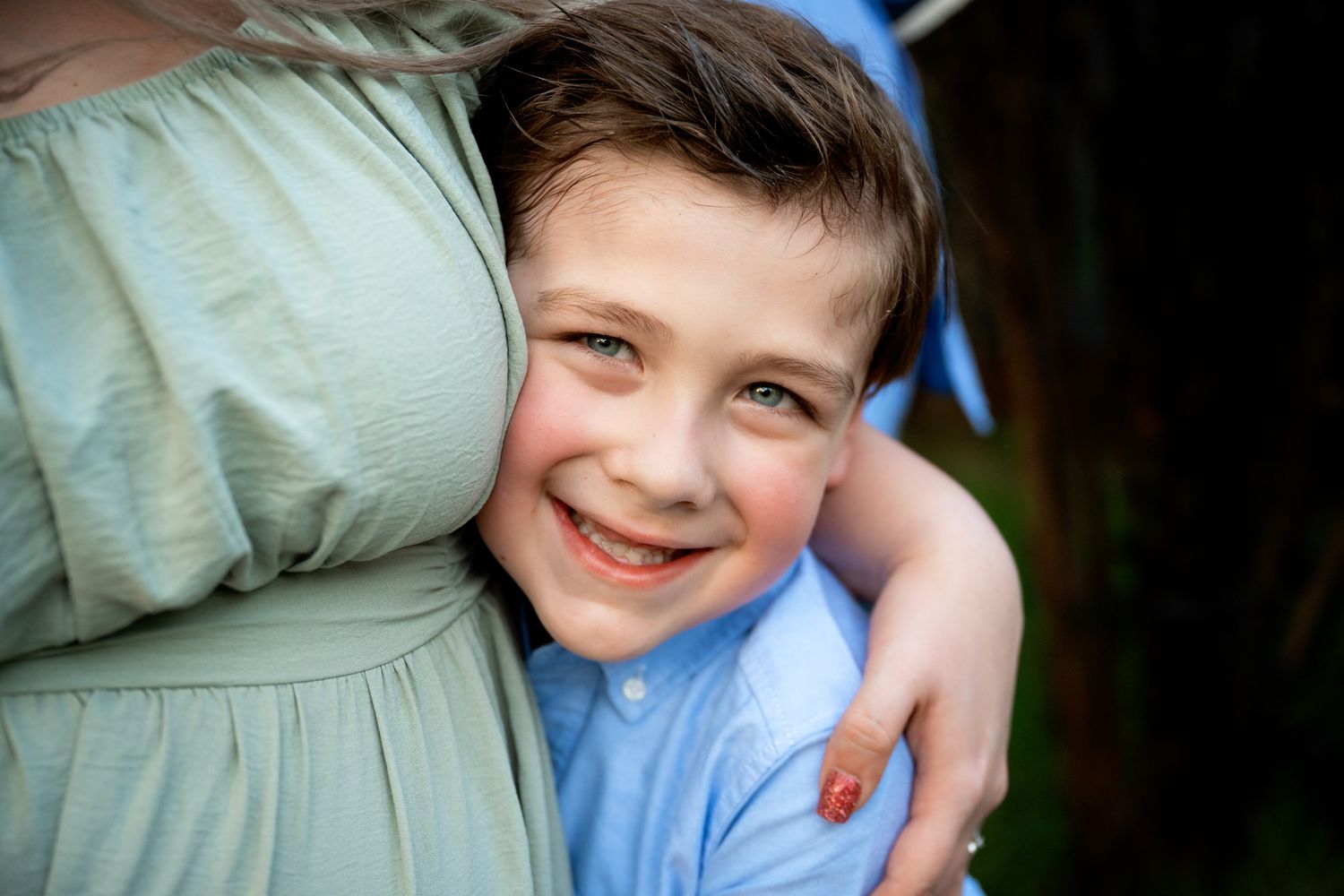 Smiling young boy hugging adult in green dress in close-up outdoor portrait.