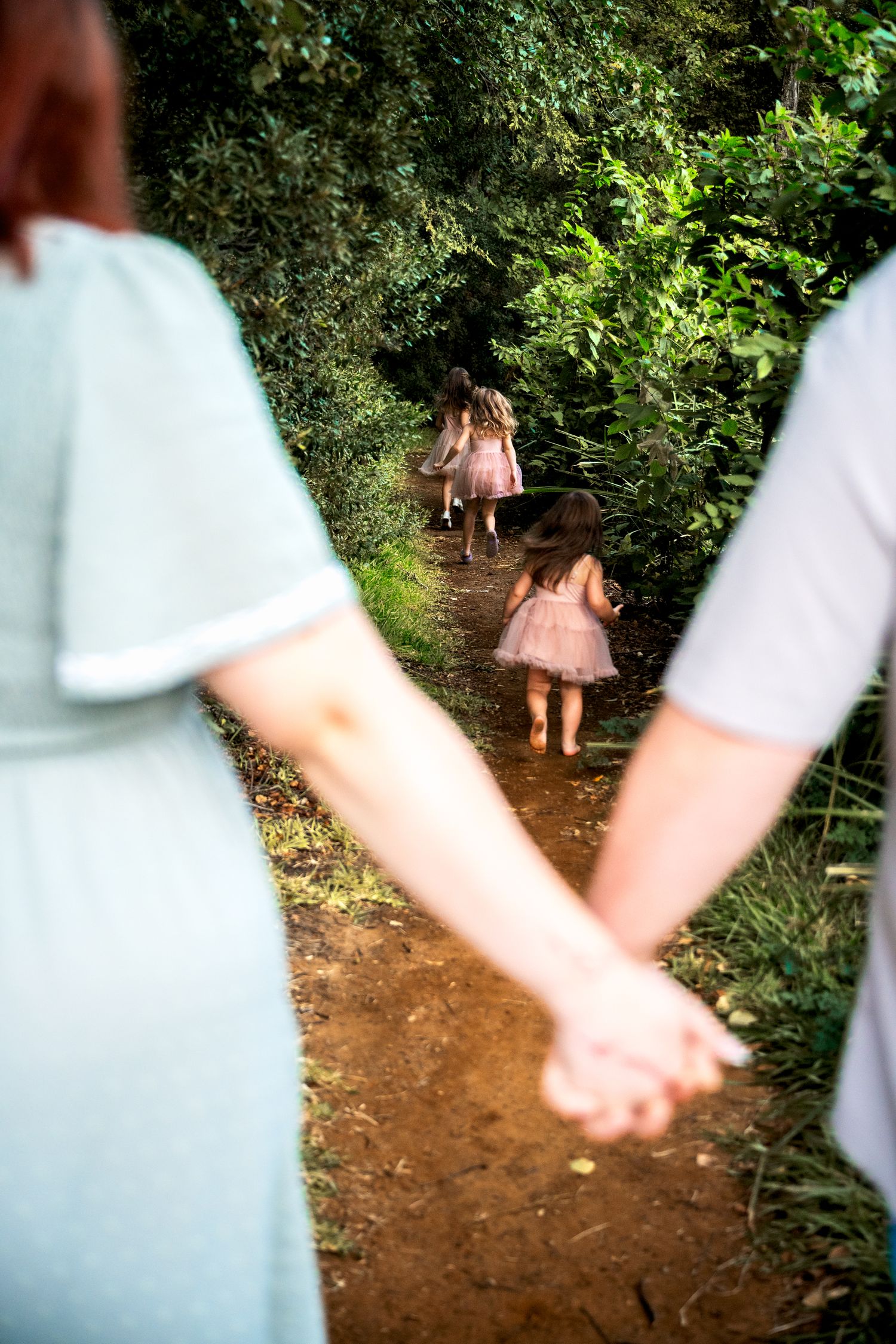 Parents hold hands watching three daughters run down a lush forest path.