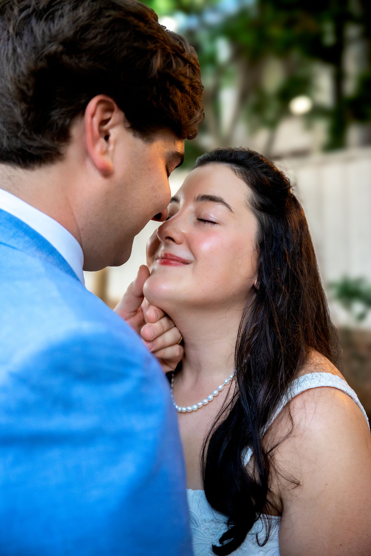 A groom in a blue suit tenderly holds a smiling bride's face as they share a romantic moment outdoors.