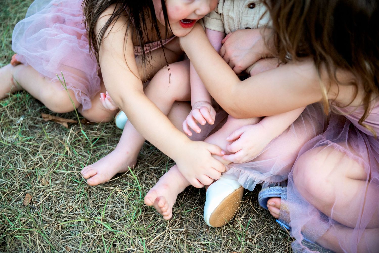 Three young girls in pink tulle dresses sit together on grass.