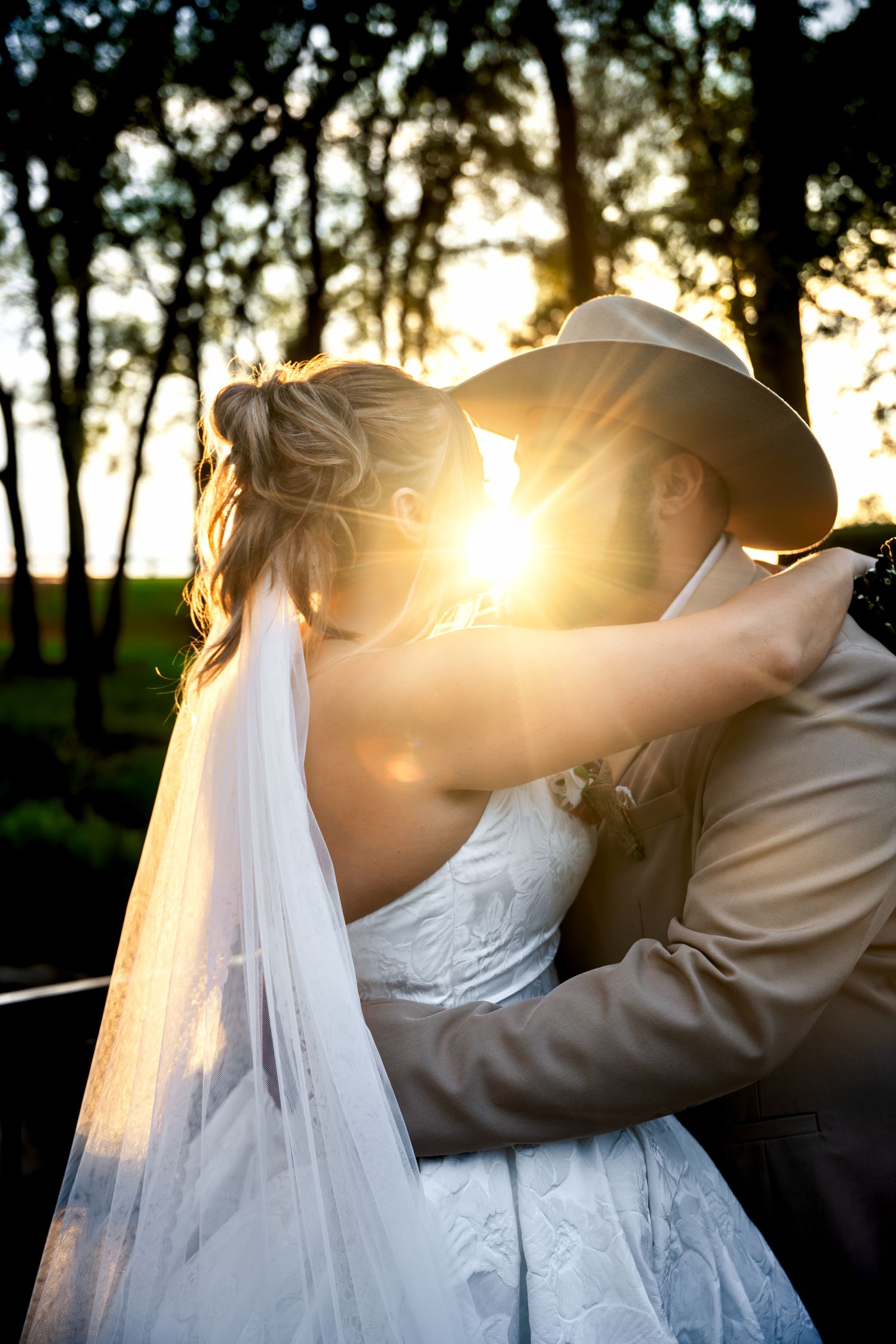Bride and groom share a romantic sunset kiss outdoors, with golden light glowing through trees, veil flowing gracefully.