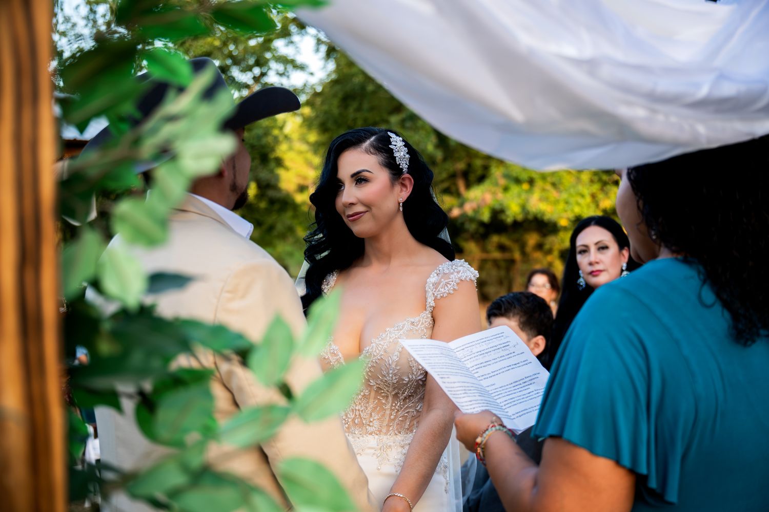 Bride smiling joyfully during outdoor wedding ceremony under floral canopy.