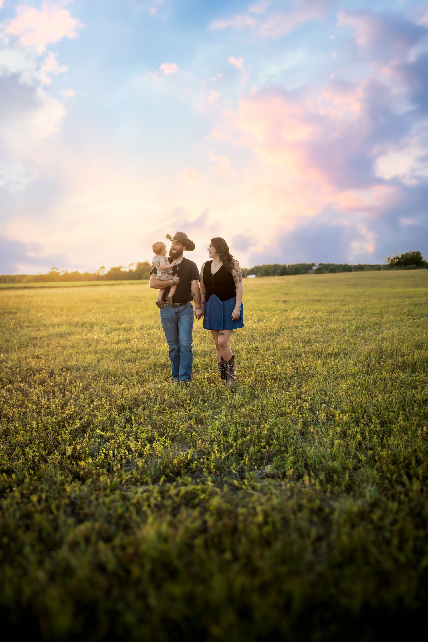 A family of three walks through a lush green field at sunset, with the father holding a baby and wearing a cowboy hat.