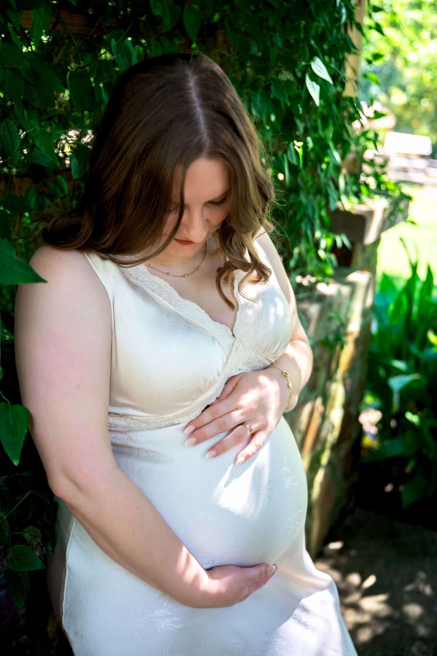 Pregnant woman in white dress cradling her baby bump in a lush green garden setting.