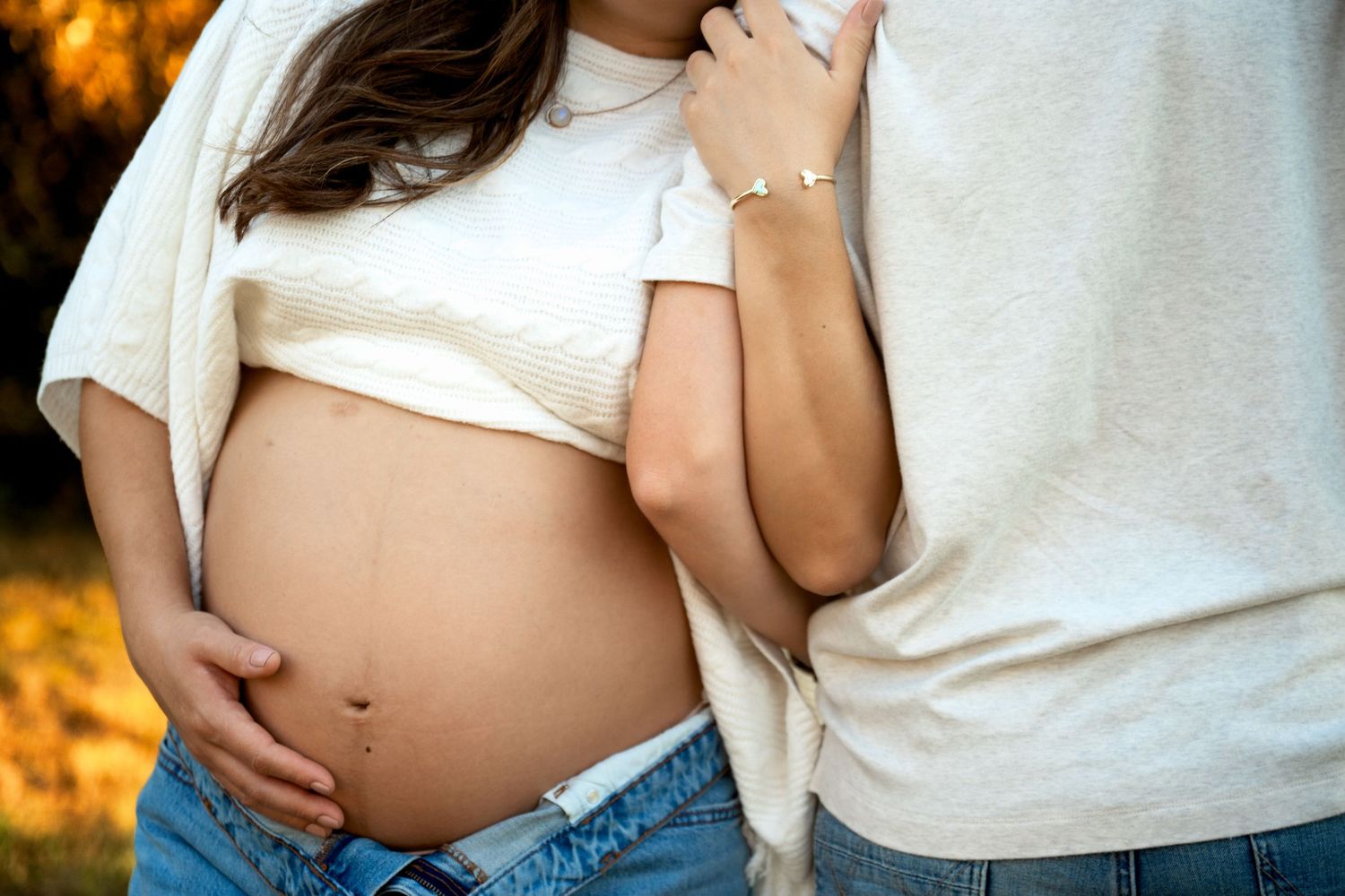 Pregnant woman in white crop top and jeans embraced by partner outdoors in warm autumn light.